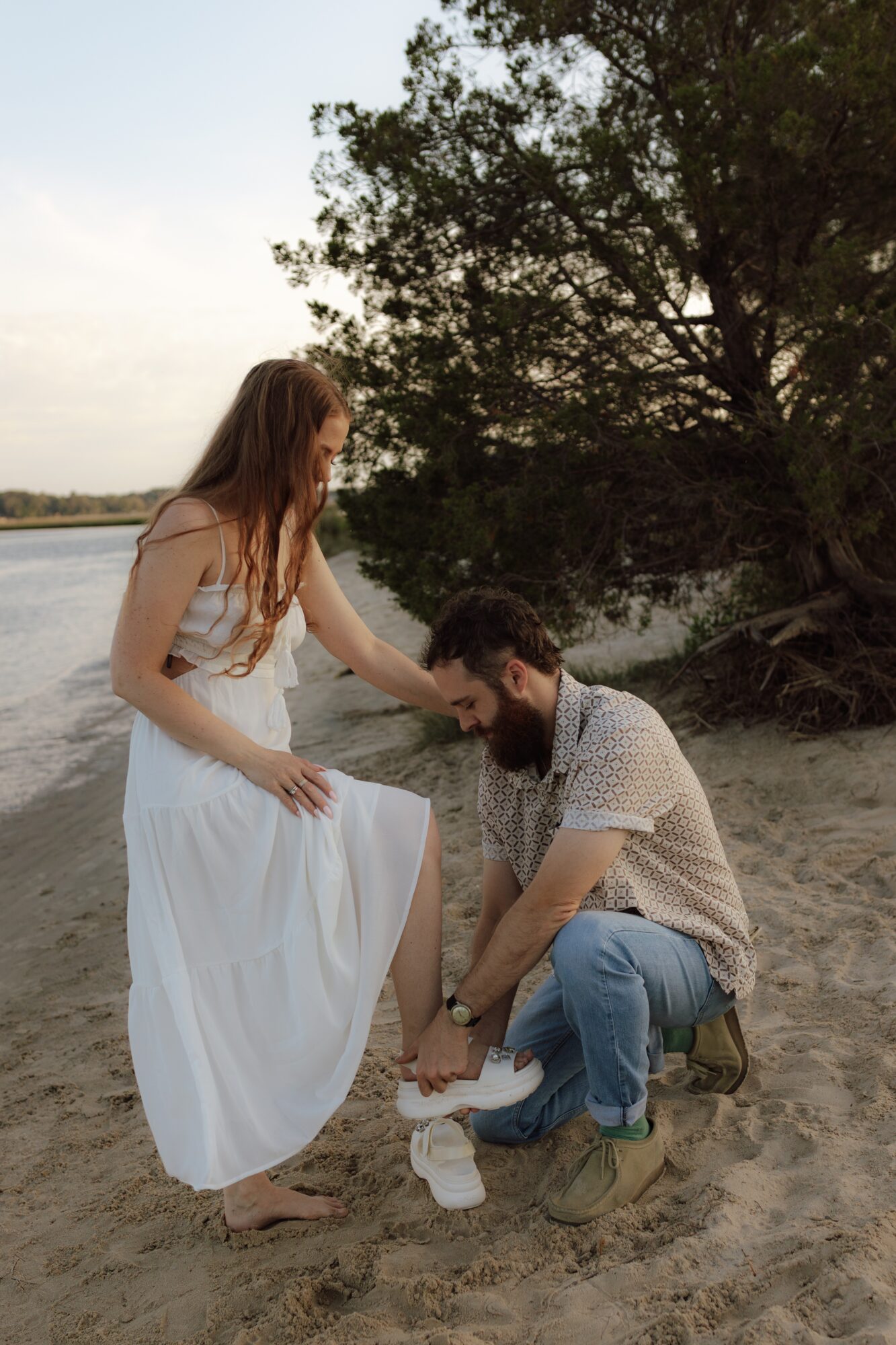 A woman in a white dress and a man with a beard are on a sandy beach near water, with trees in the background.