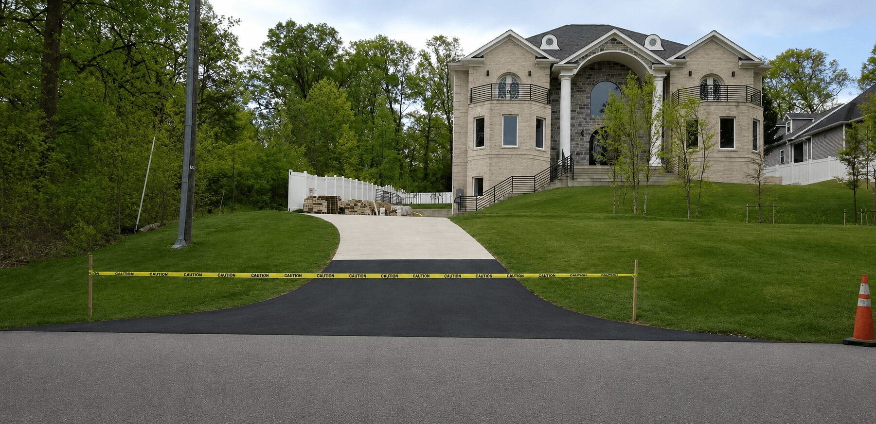 A large house with a curved driveway, surrounded by green grass and trees, with a yellow barrier and orange cone.