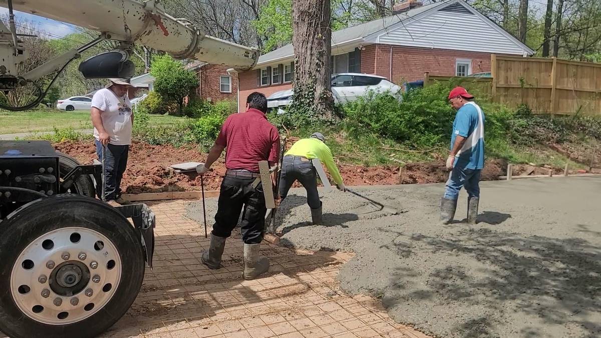 Four people working on a sidewalk construction project outdoors, with a tree, houses, and a parked car in the background.