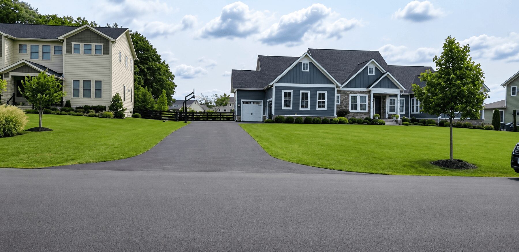 Residential neighborhood with two houses, green lawns, trees, and a paved street under a partly cloudy sky.