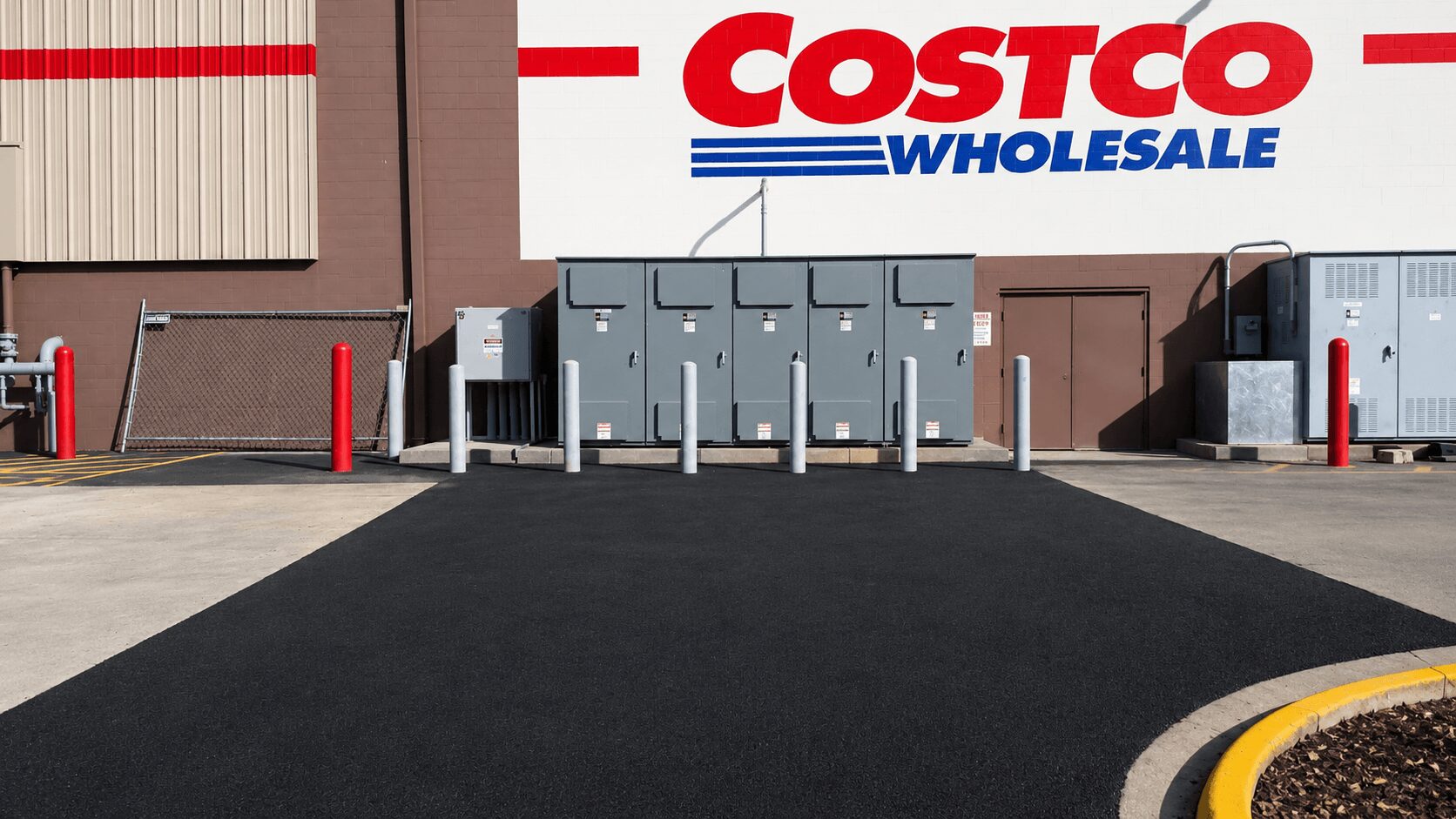 View of a Costco Wholesale store's loading area with electrical boxes and red bollards, and a black asphalt surface.