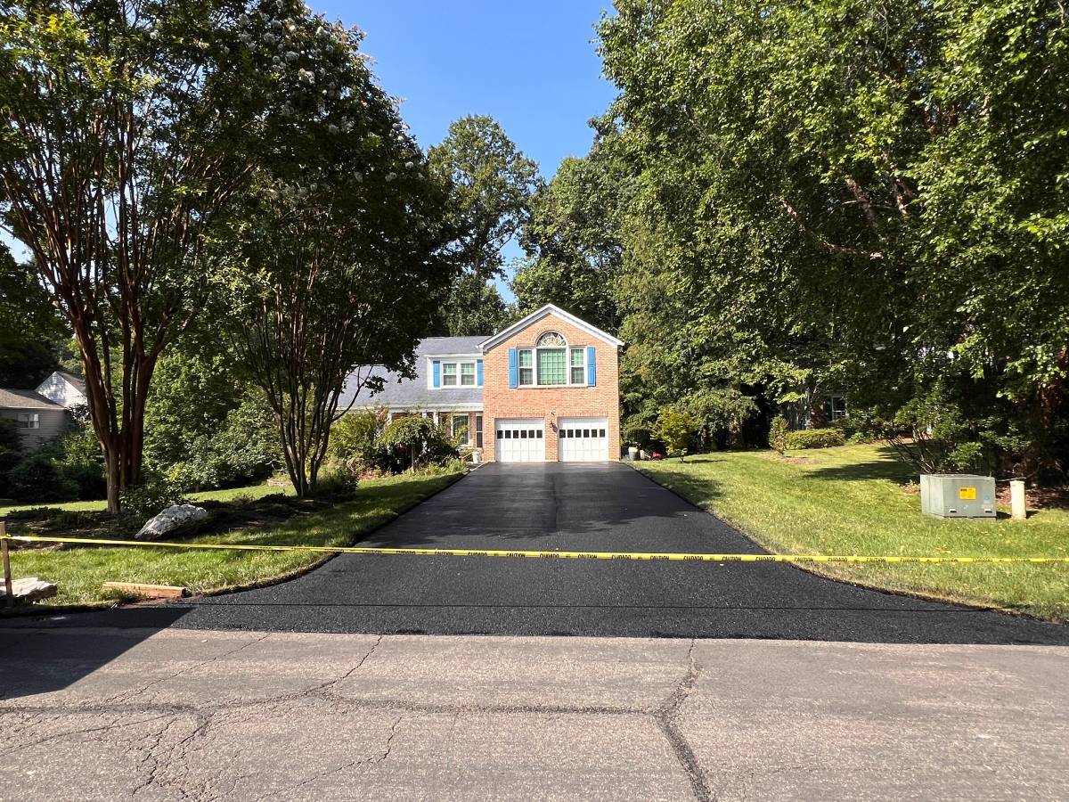 A house with a driveway, surrounded by trees and greenery, under a clear blue sky.