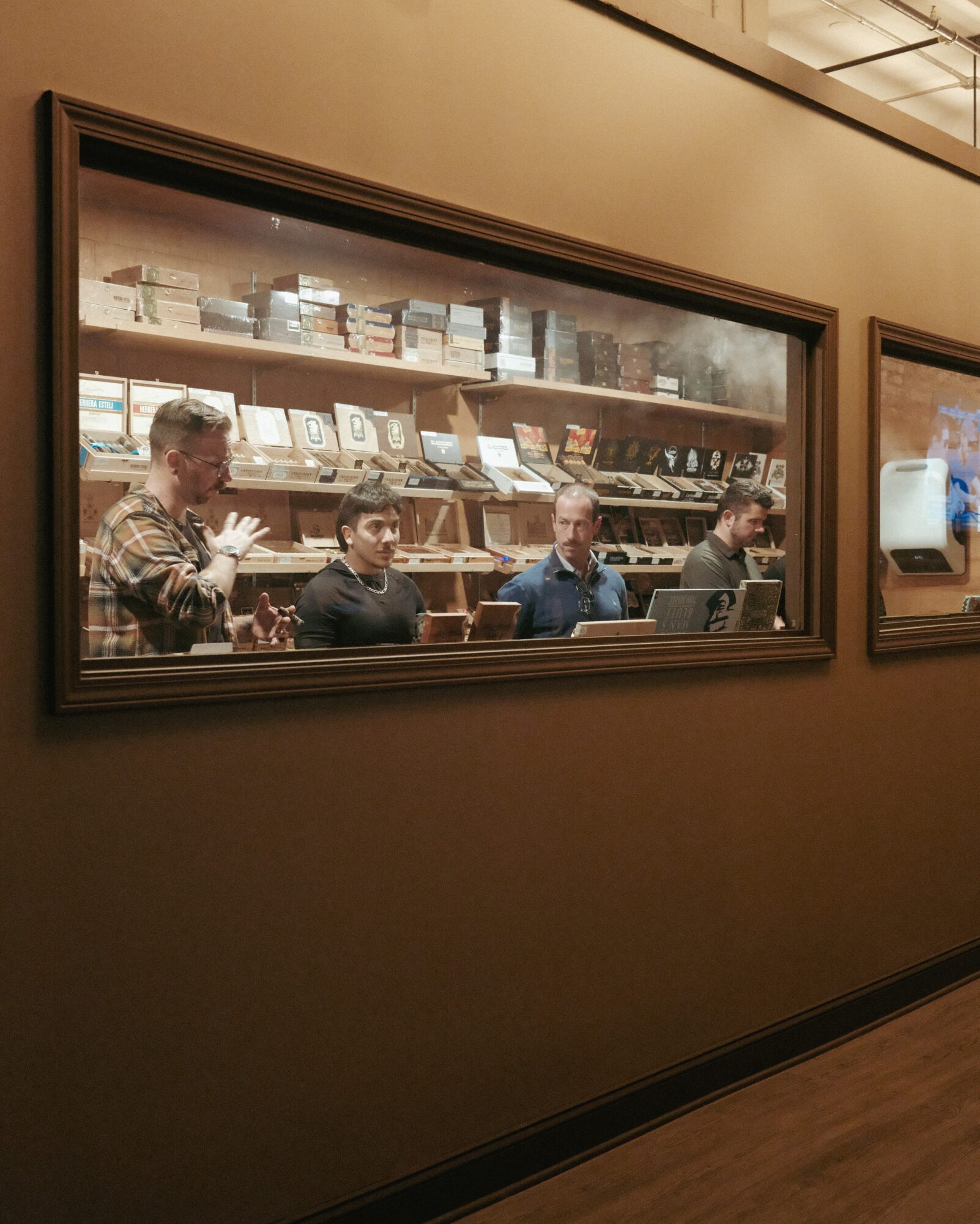 Four people sit at a table inside a room with shelves of books or items behind them, viewed through a large window.