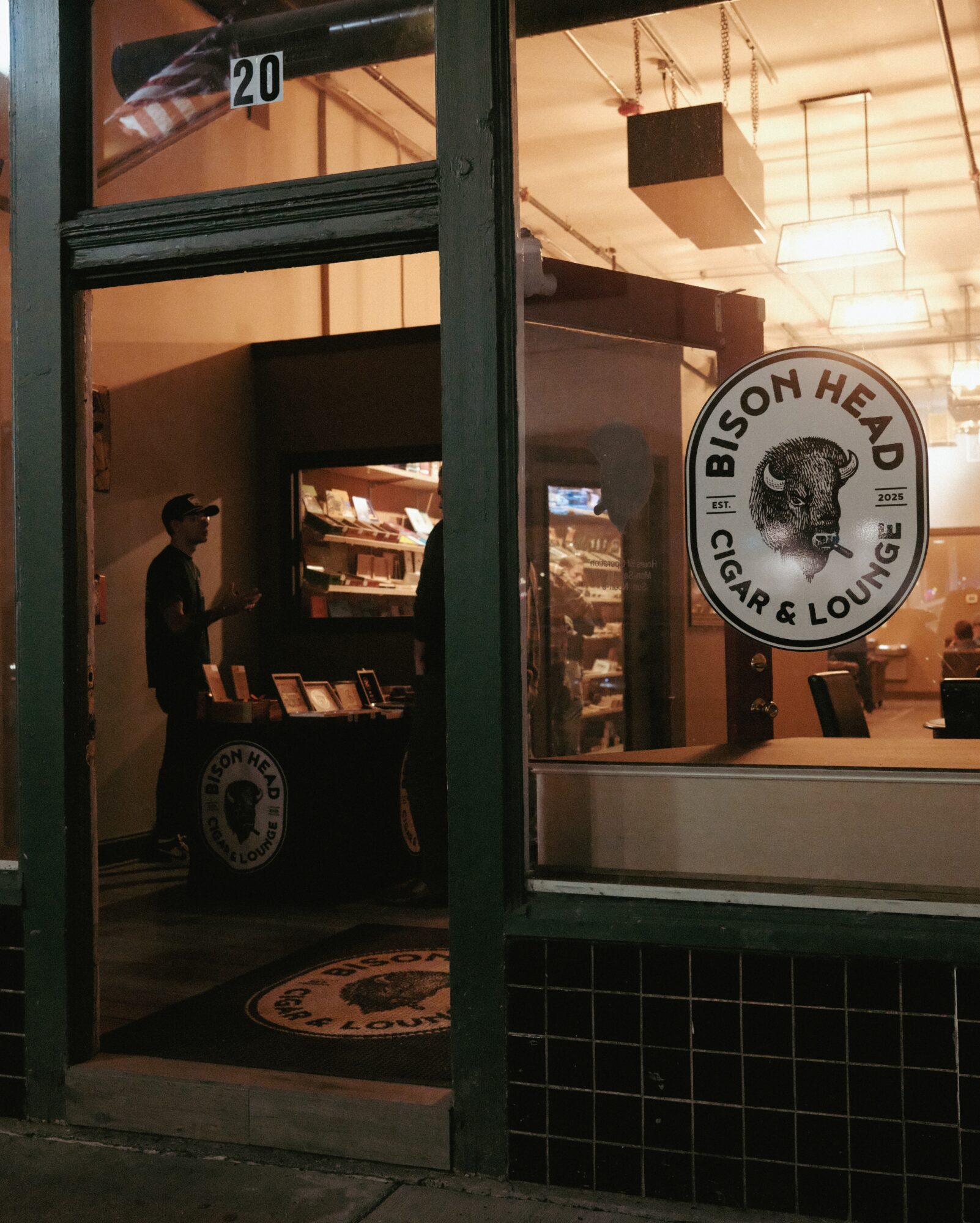 View through a glass door into a cigar lounge with a circular sign, a person standing, and shelves with items inside.