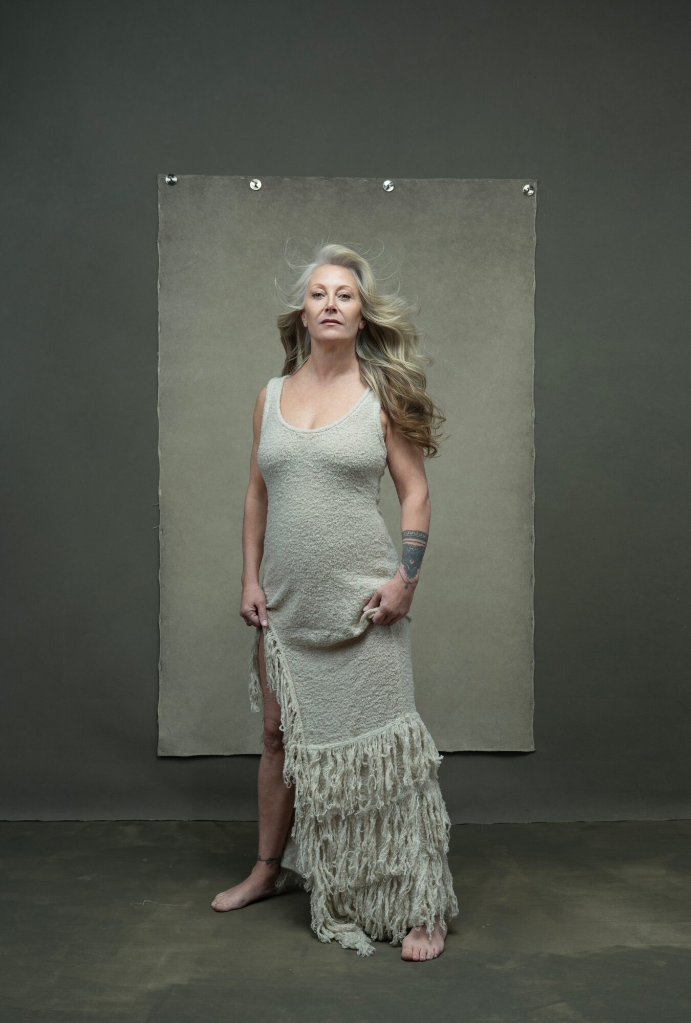 Woman with long hair in a sleeveless dress standing in front of a gray backdrop, hands at sides, looking at camera.