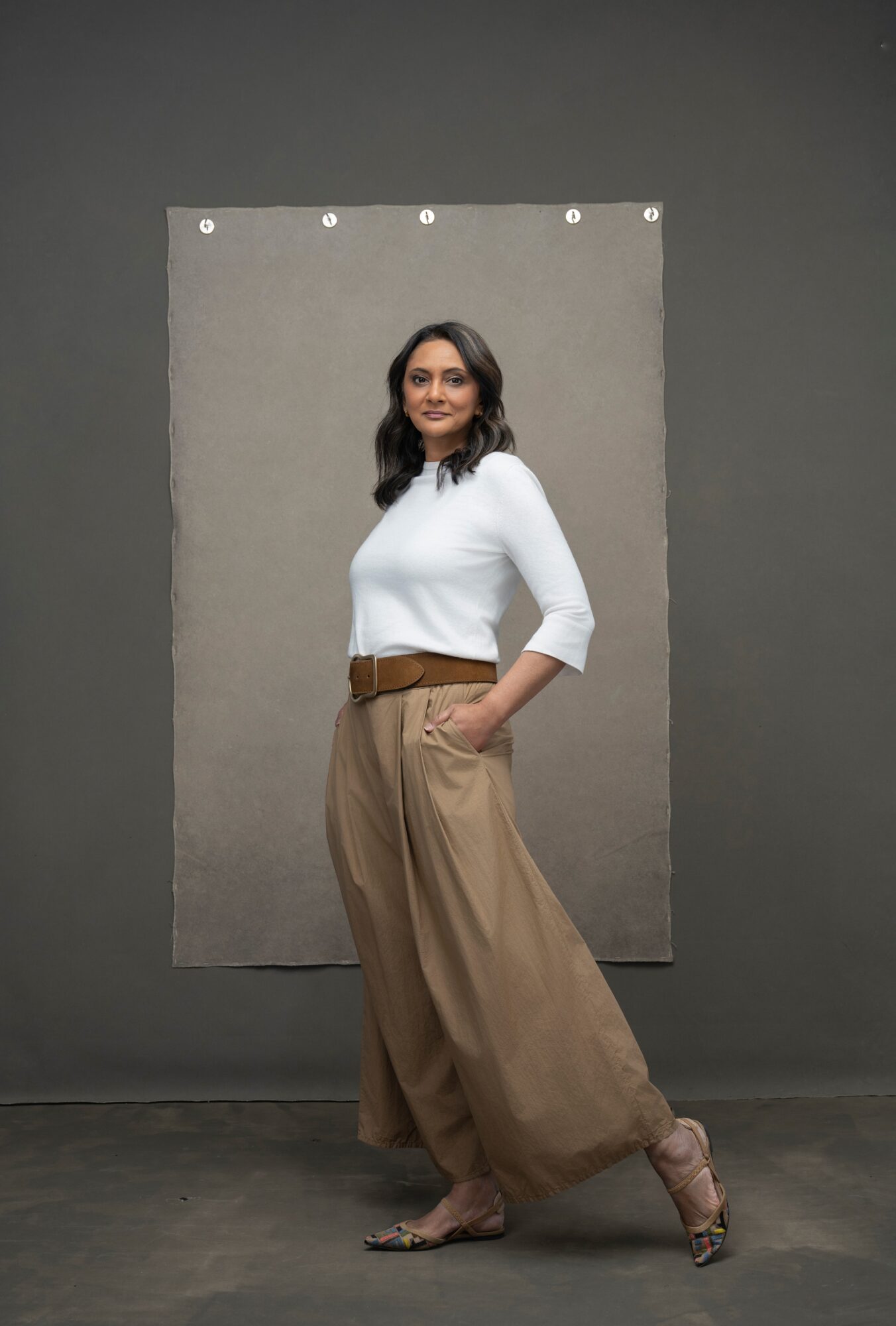 Woman standing with hands in pockets, wearing white top and wide brown pants, against a gray background.