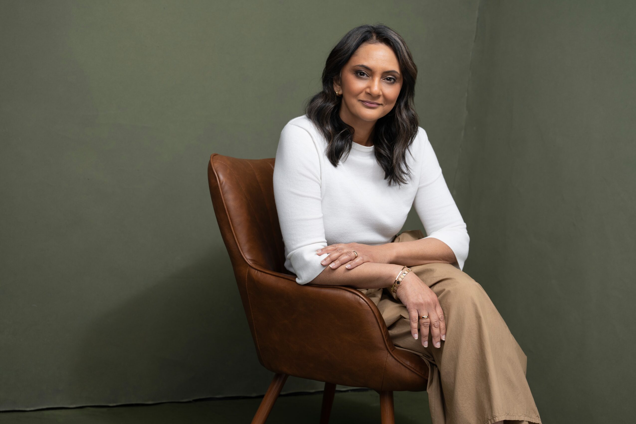 Woman with dark hair sitting on a brown chair against a green wall, smiling, wearing a white top and beige pants.