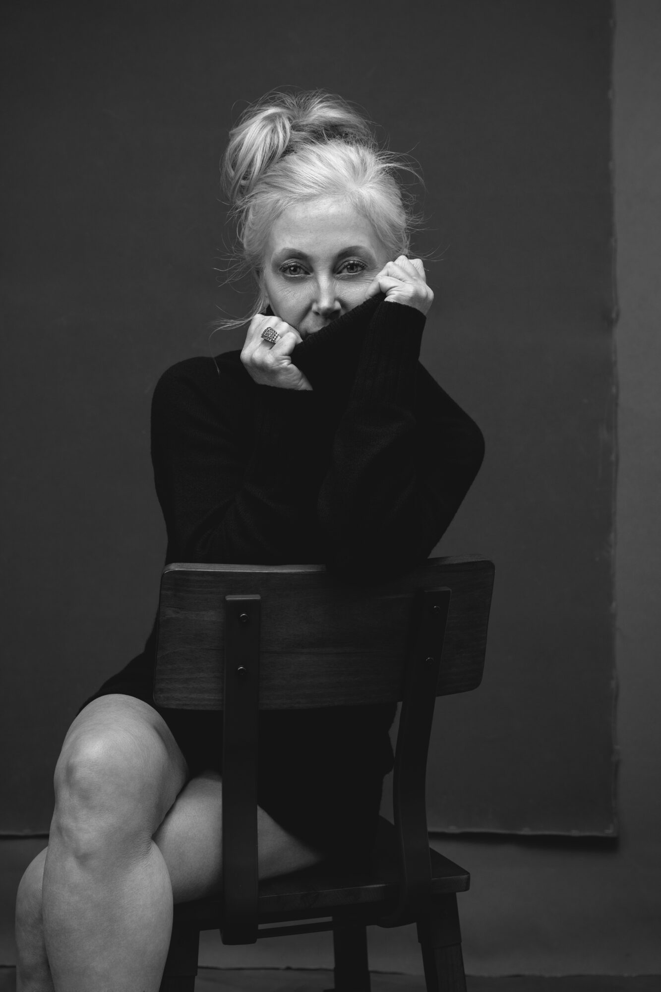 Young woman with light hair in a messy bun, sitting on a chair, covering her mouth with her hands, wearing a dark sweater, in black and white.