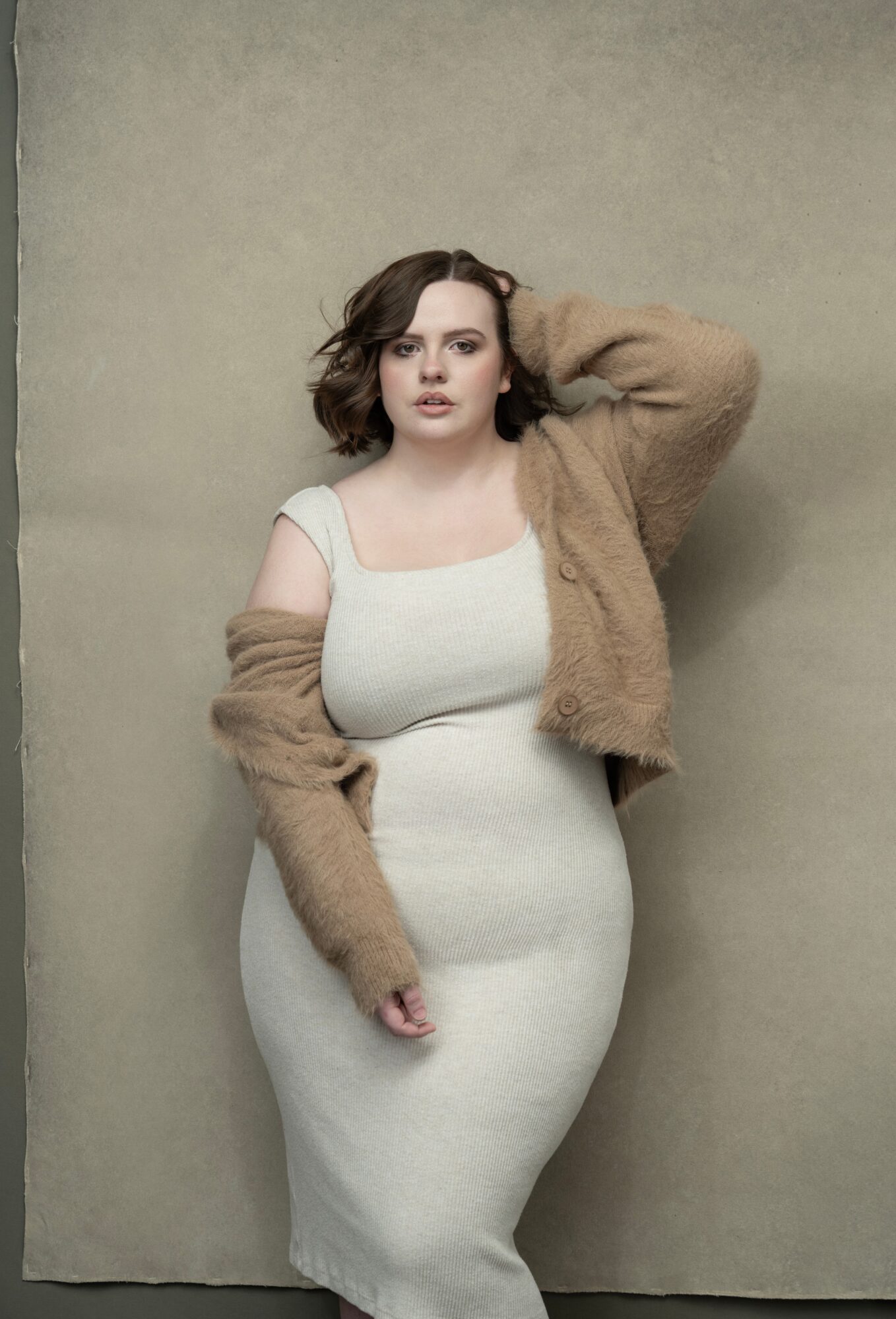 Woman with short wavy hair wearing a white dress and a brown fuzzy jacket, standing against a plain background.
