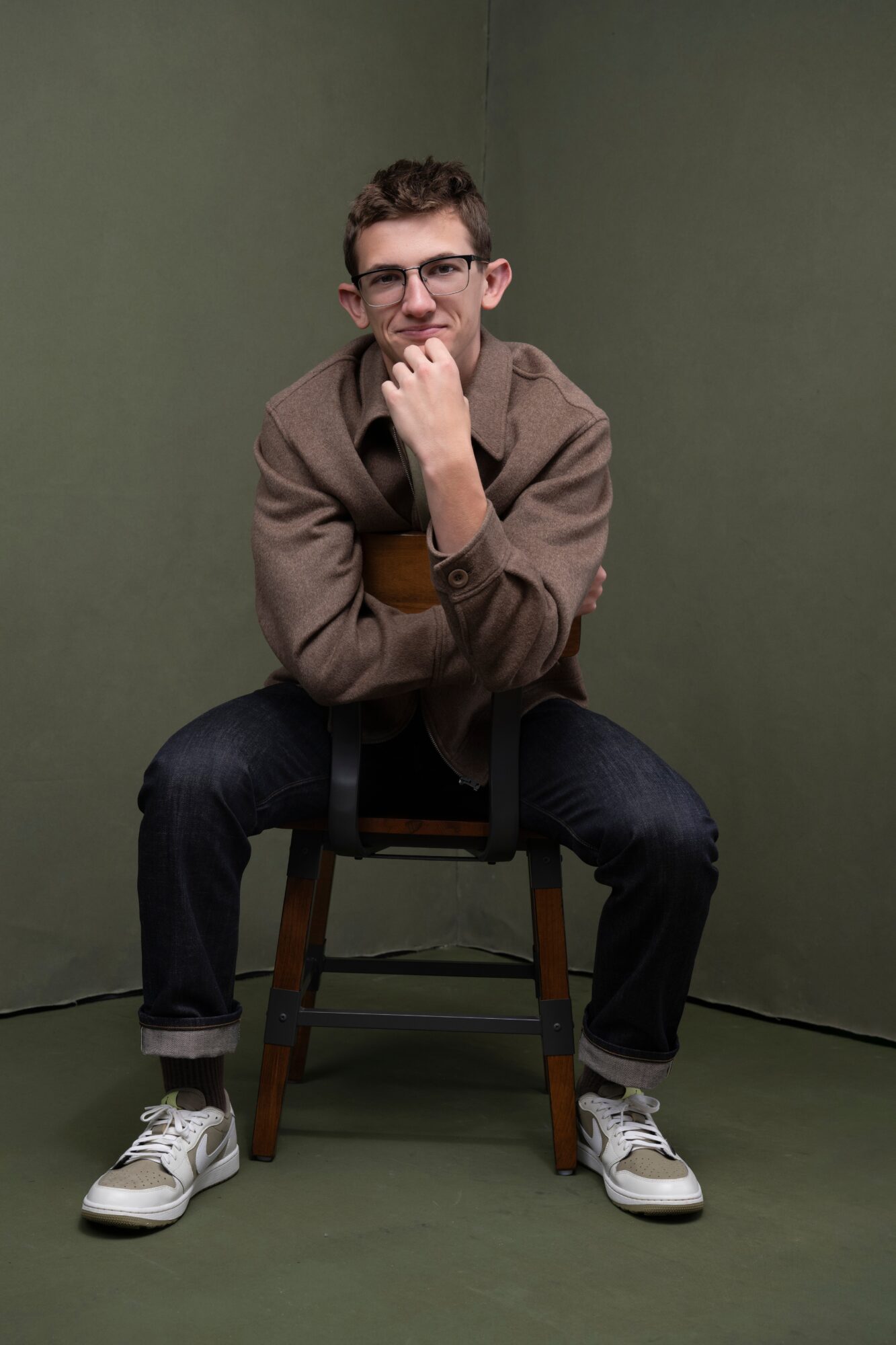 Young man sitting on a chair with a green background, wearing glasses, a brown jacket, and sneakers, smiling with hand on chin.