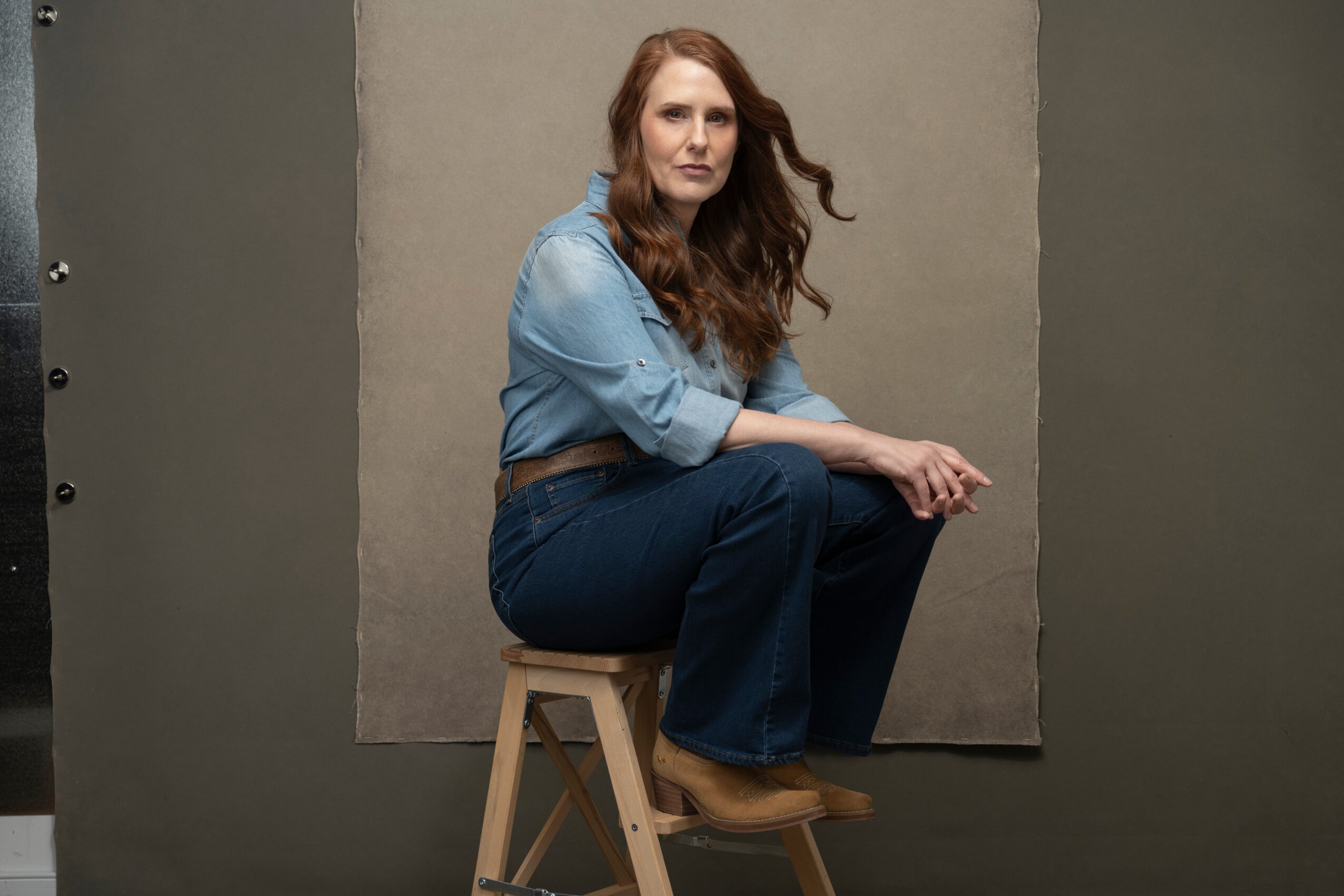 Woman with long wavy hair sitting on a stool against a plain background, wearing a denim shirt and jeans.