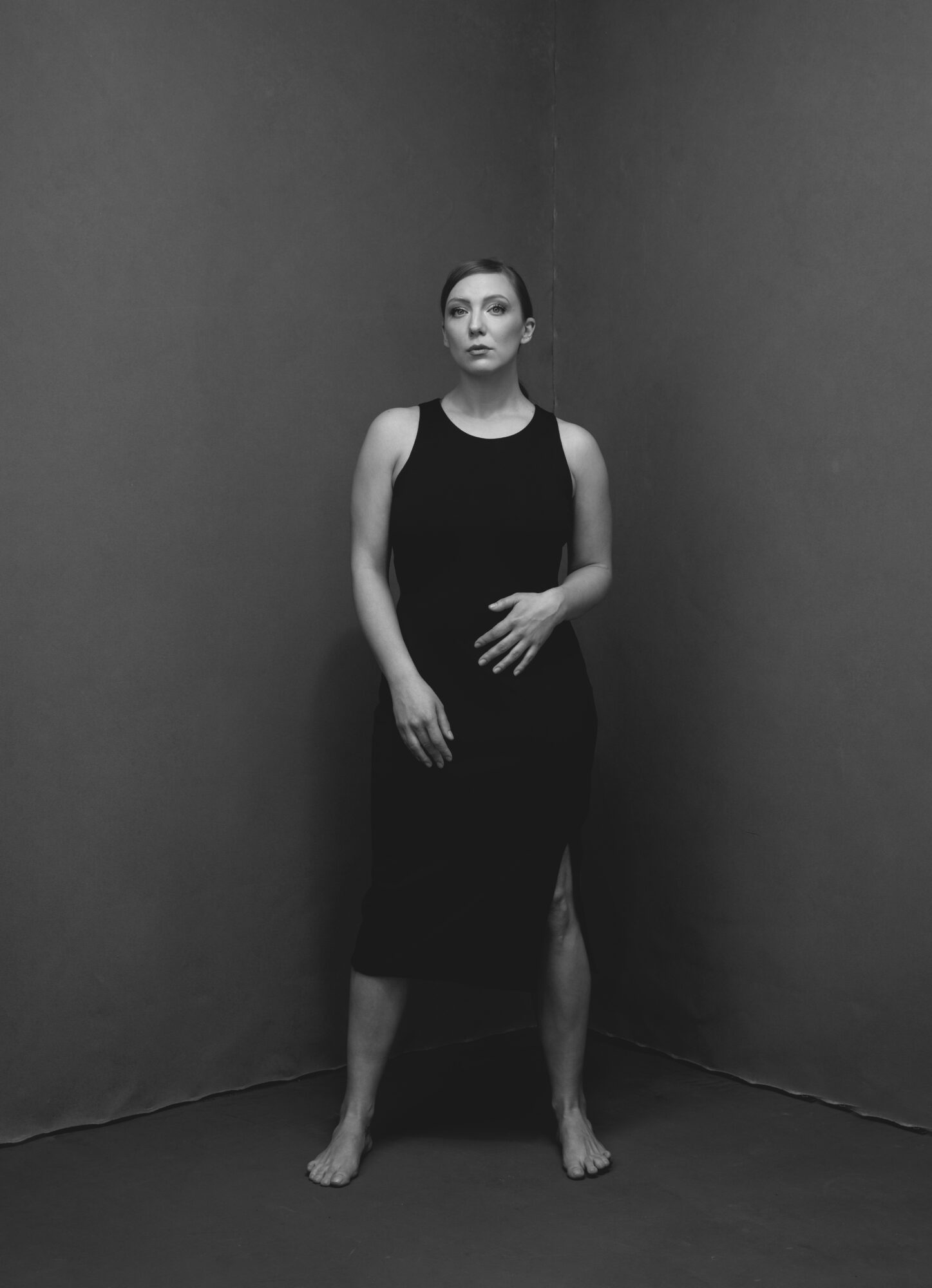 Woman standing barefoot in a corner, wearing a sleeveless black dress, with hands resting on her stomach, looking at the camera.