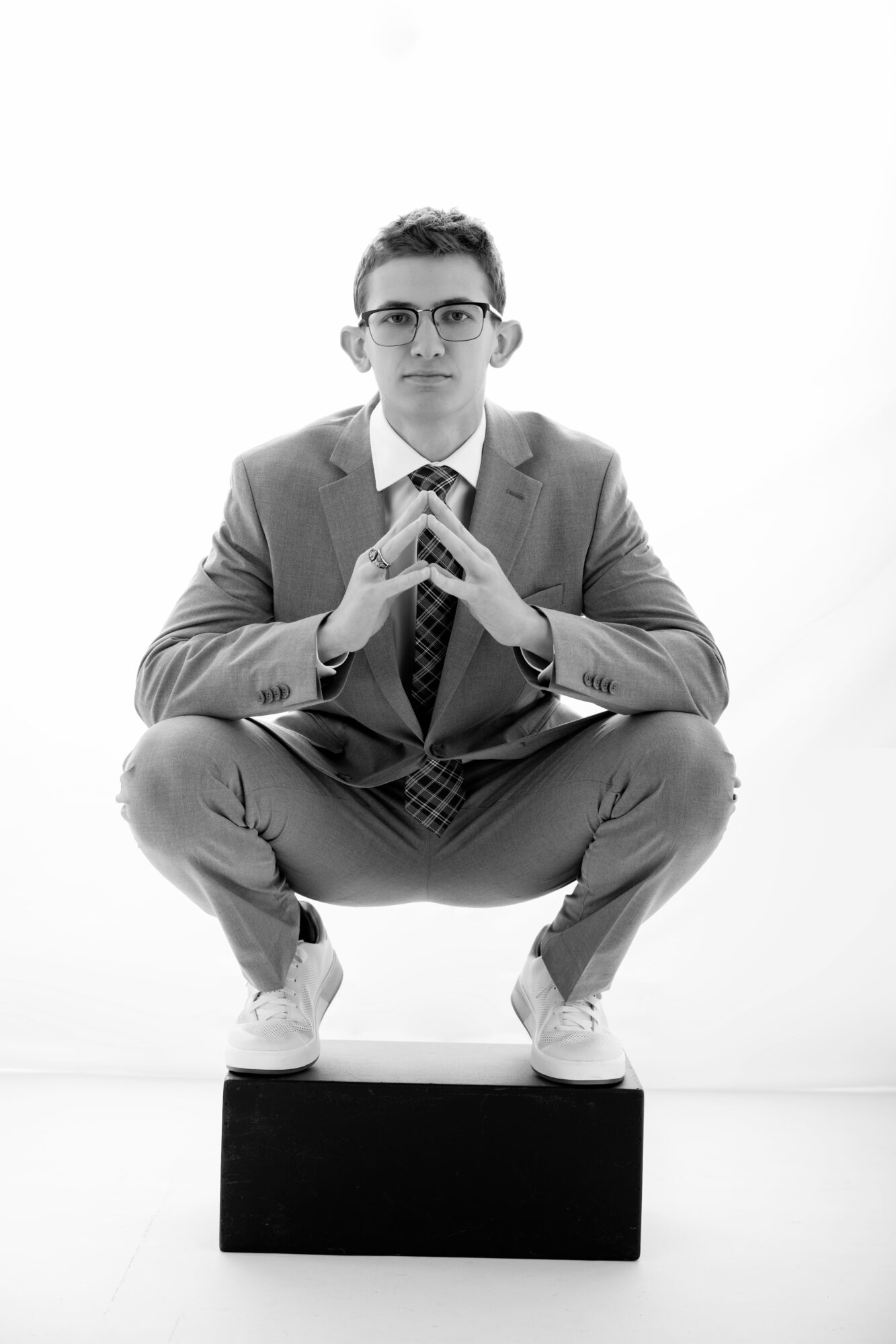 Young man in suit squatting on a black block, hands clasped, looking at camera, white background, glasses, formal attire.