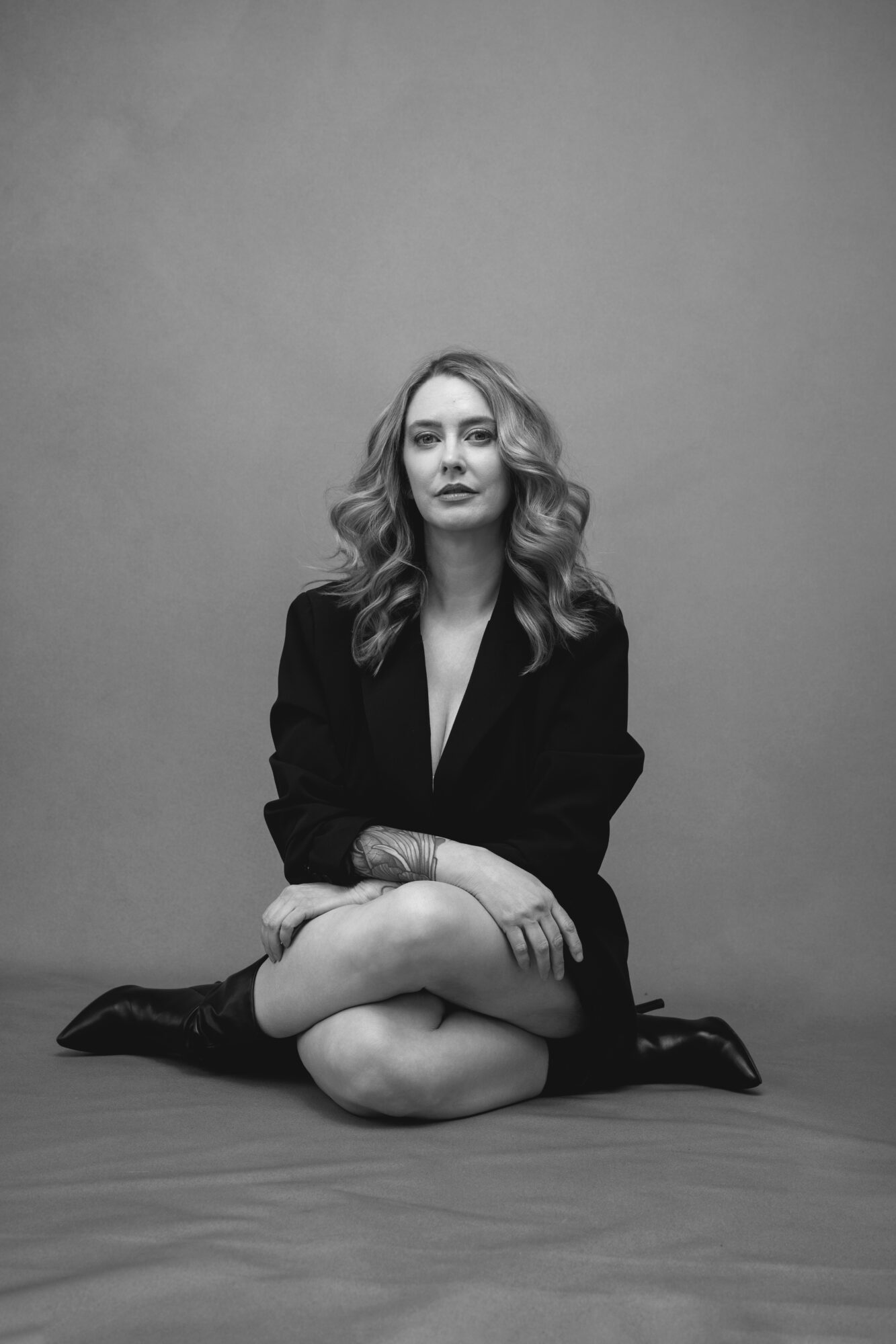 Woman sitting cross-legged on the floor, wearing a blazer, with wavy hair, against a plain background.