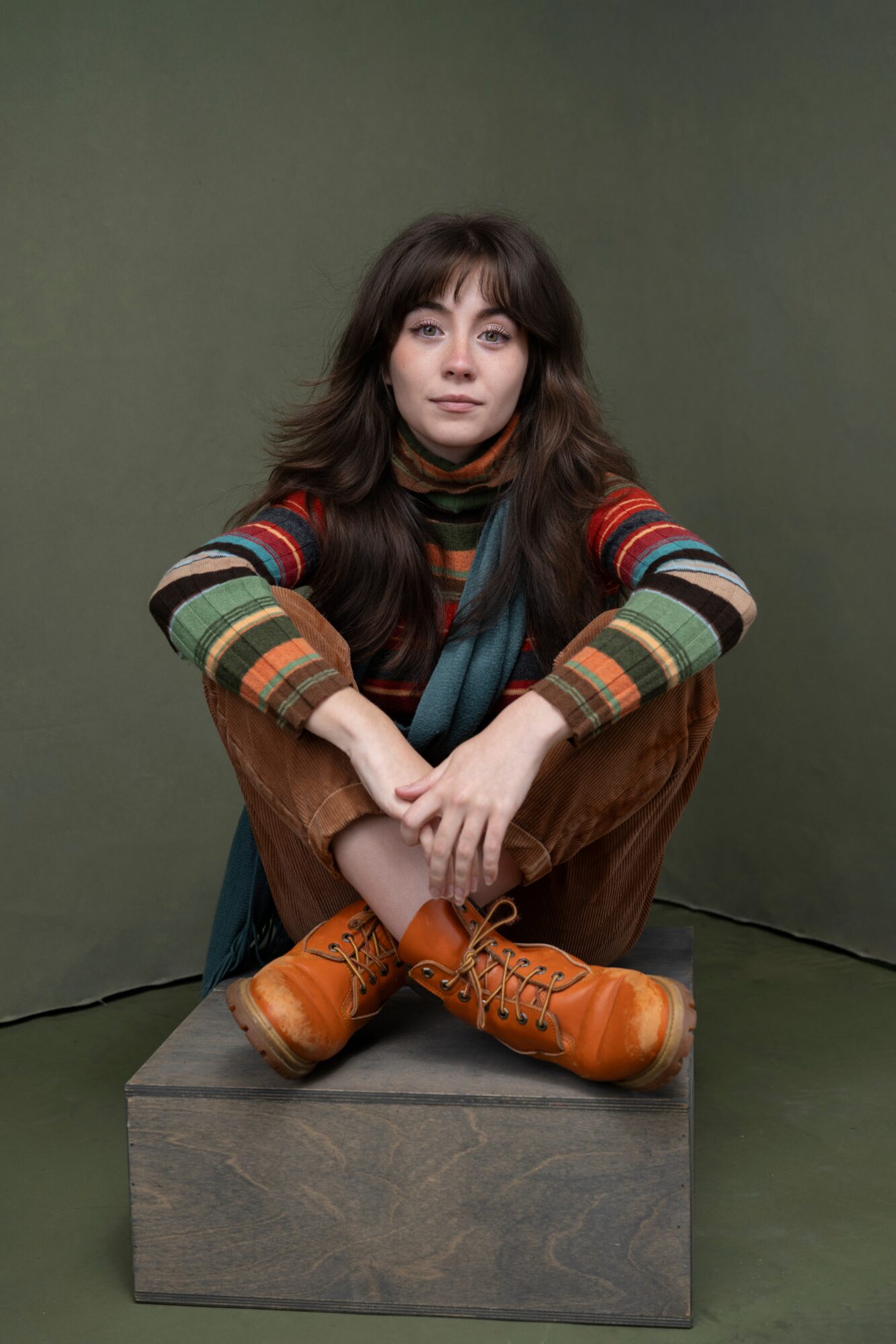 Young woman with long dark hair sitting cross-legged on a wooden block, wearing a colorful striped sweater and brown pants, against a plain green background.