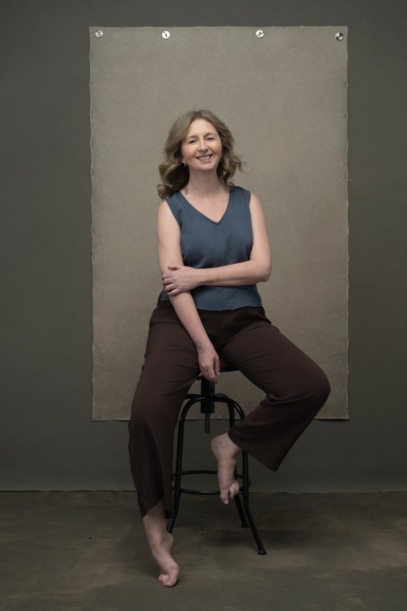 Woman with shoulder-length hair smiling, sitting on a stool with crossed arms, in front of a gray backdrop.