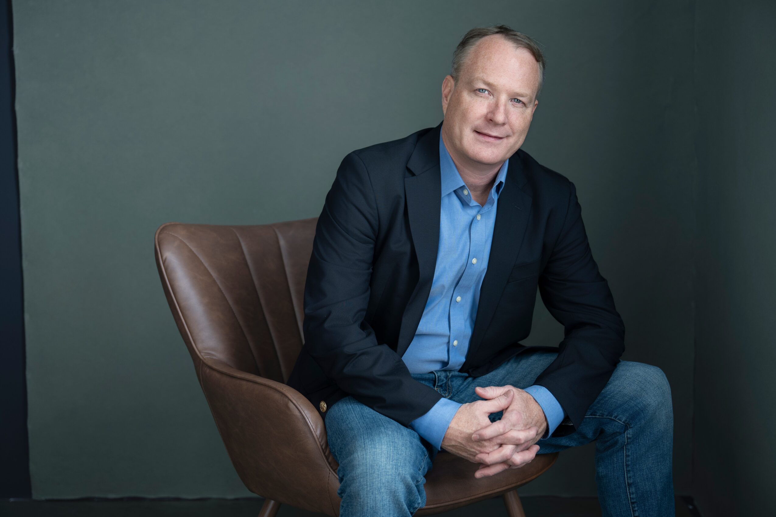 Man sitting on a brown chair, wearing a dark blazer and blue shirt, smiling, against a plain dark background.