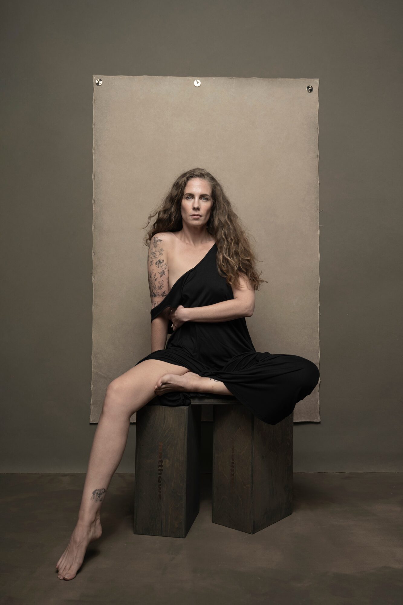 Woman with long curly hair in a black dress sitting on a wooden block, with a plain background behind her.