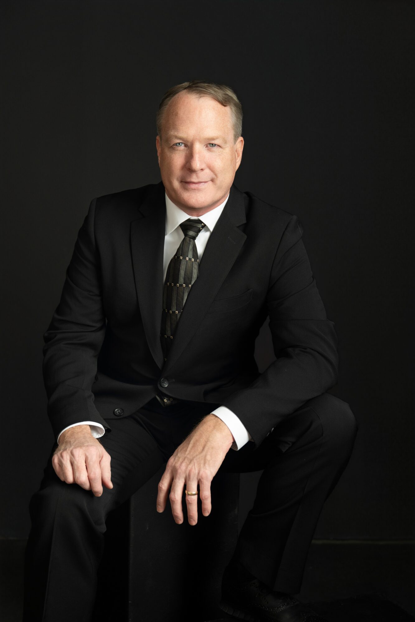 Man in a black suit and tie sitting against a dark background, looking at the camera.
