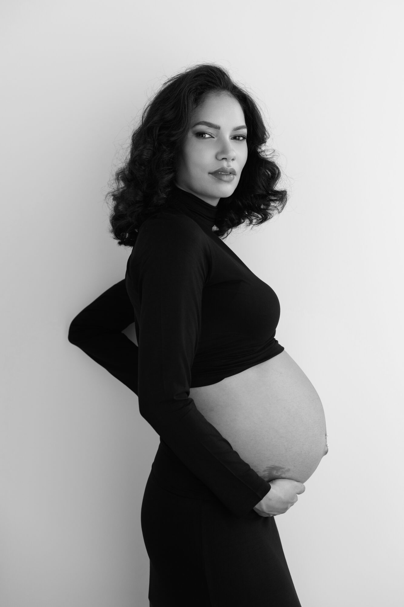 Pregnant woman with curly hair in black clothing, standing sideways, hand on belly, looking at camera, black and white photo.