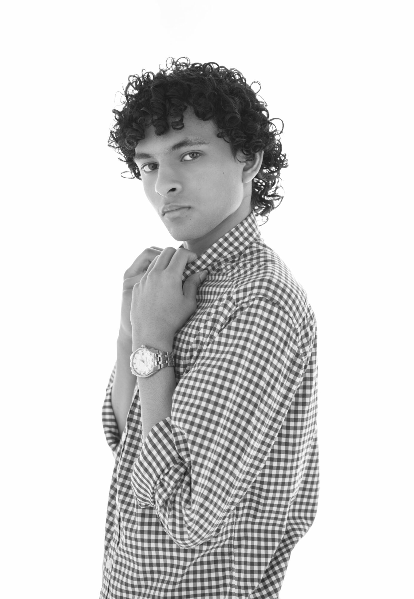 Young person with curly hair wearing checkered shirt and watch, looking at camera, hand near face, neutral expression.
