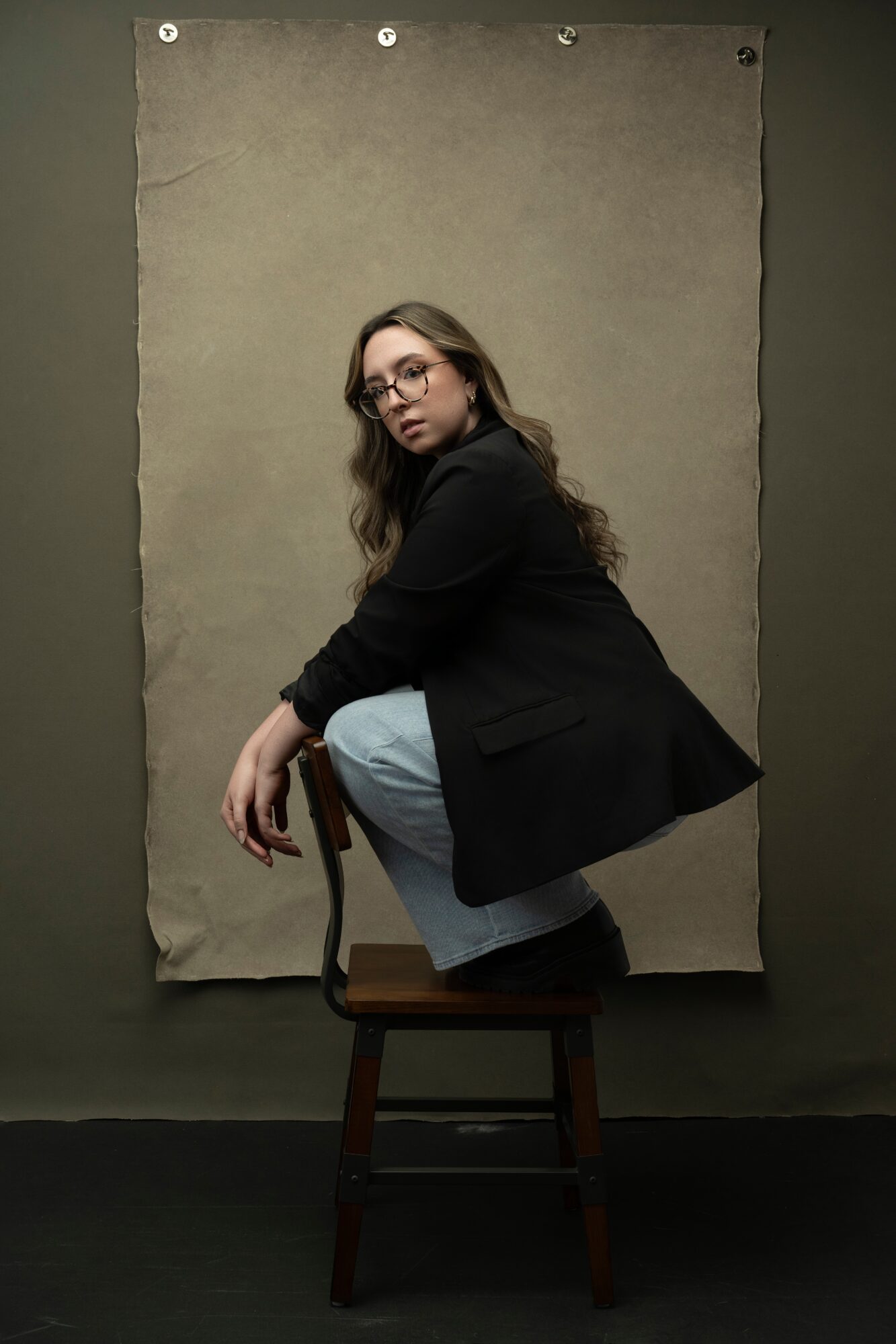 Person with long hair and glasses crouching on a chair, wearing a black coat and light pants, against a plain background.