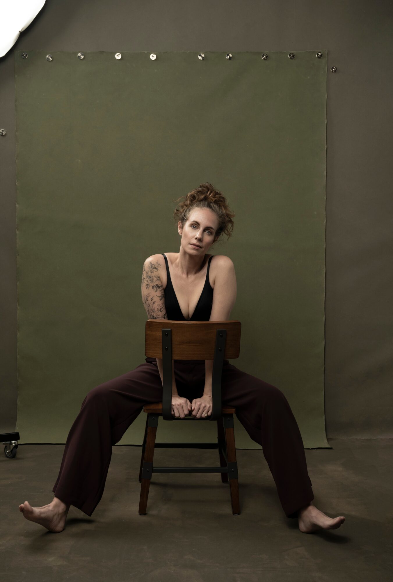 Person sitting on a chair with legs spread wide, leaning forward, in front of a green backdrop, with tattoos on arms, wearing dark clothing.