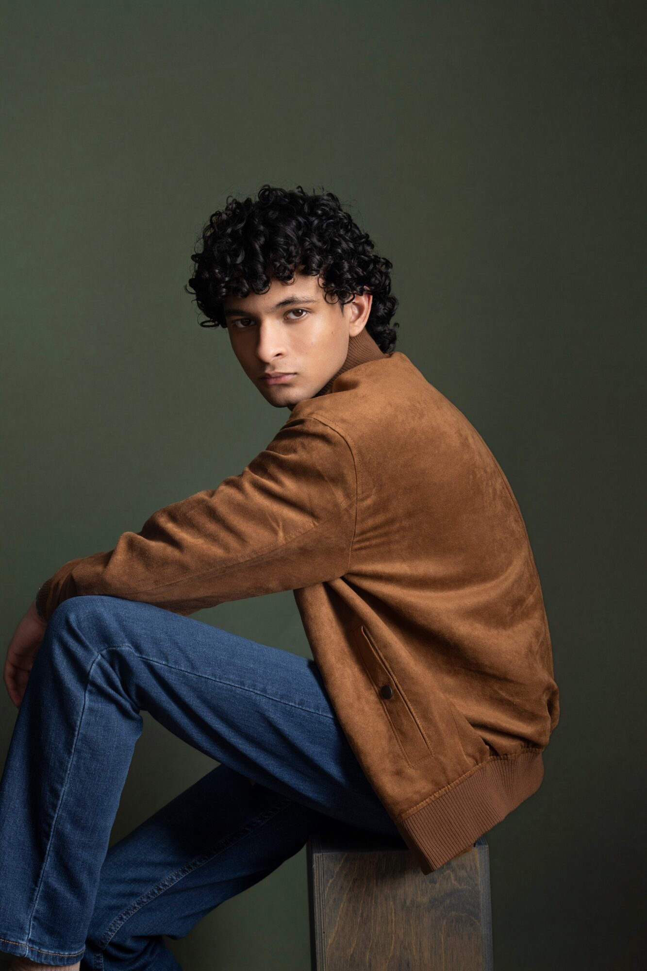 Young man with curly hair sitting on a wooden block, wearing a brown jacket and blue jeans, against a dark green background.