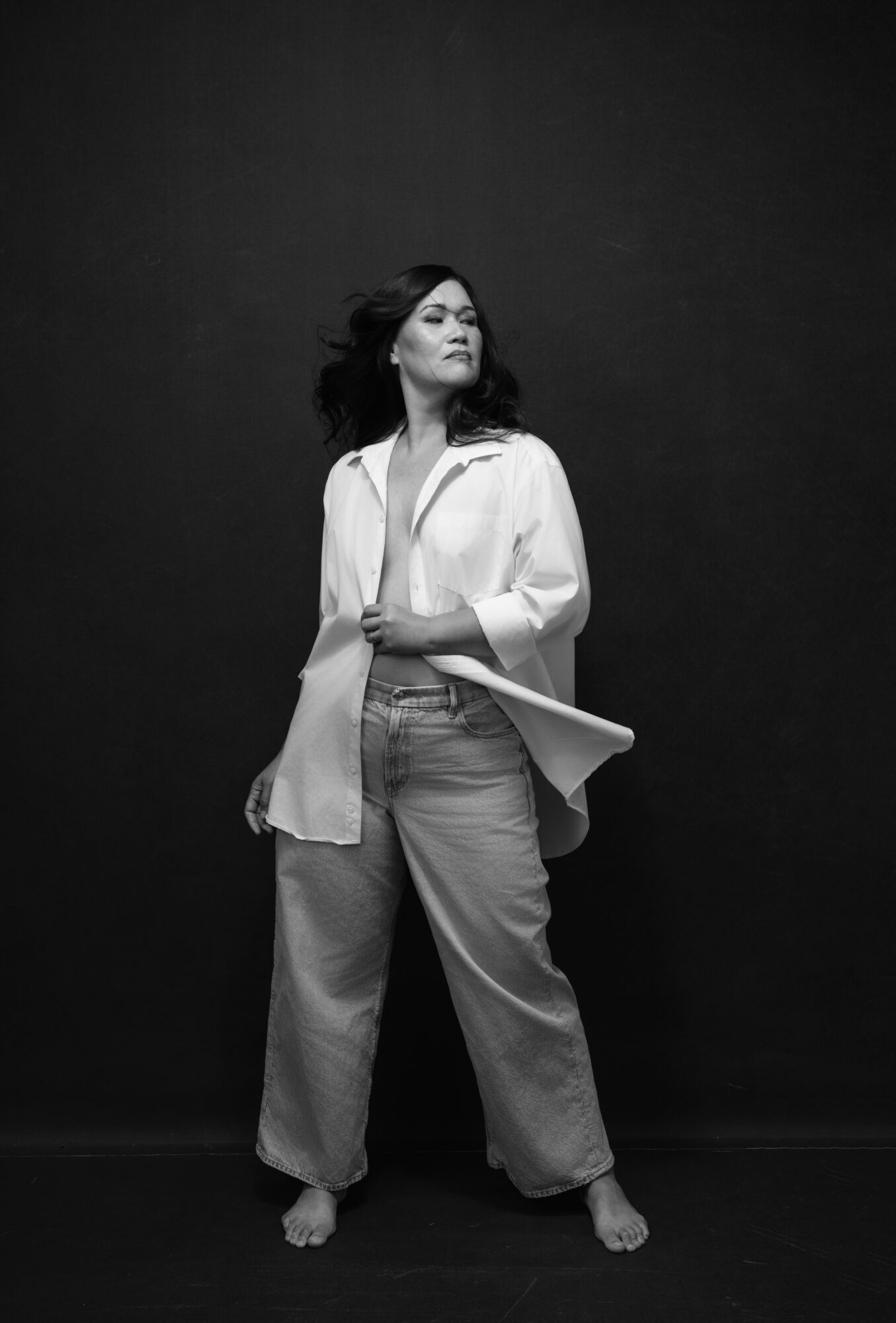 Woman standing barefoot against a dark background, wearing a white shirt and wide pants, looking to the side.