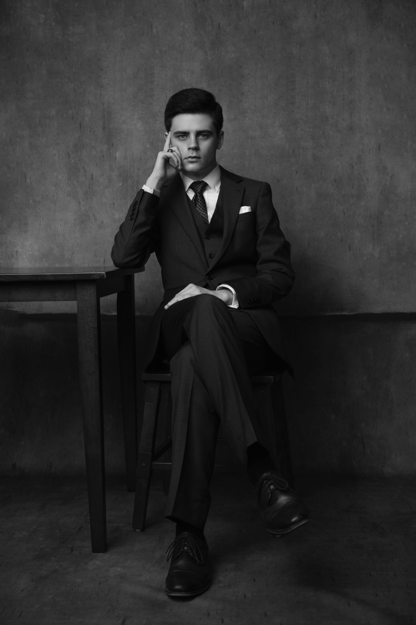 Man in suit sitting on chair, holding phone to ear, with a serious expression, against a plain wall background.