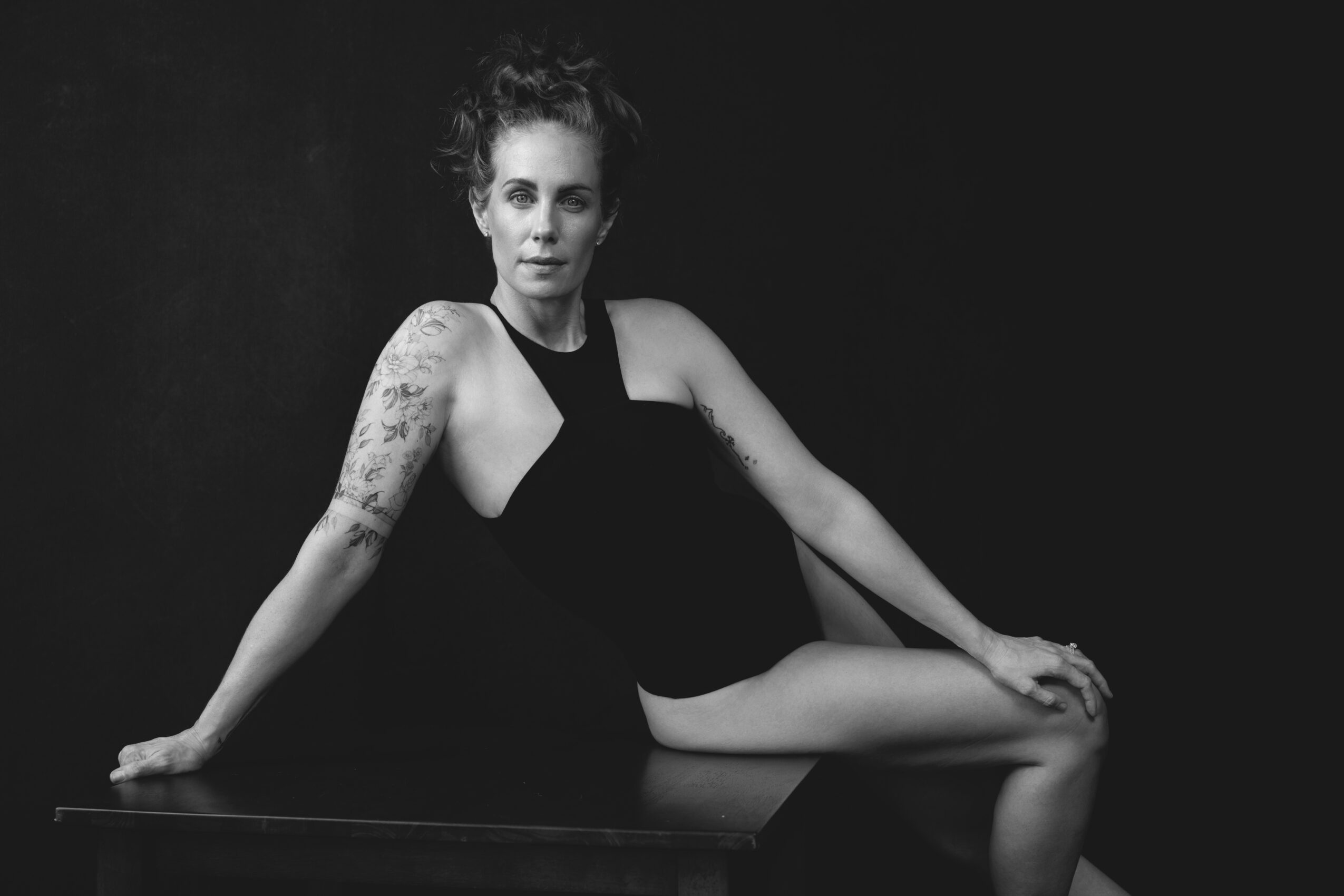 Woman in black swimsuit sitting on a surface with one arm supporting her, black background, black and white photo.