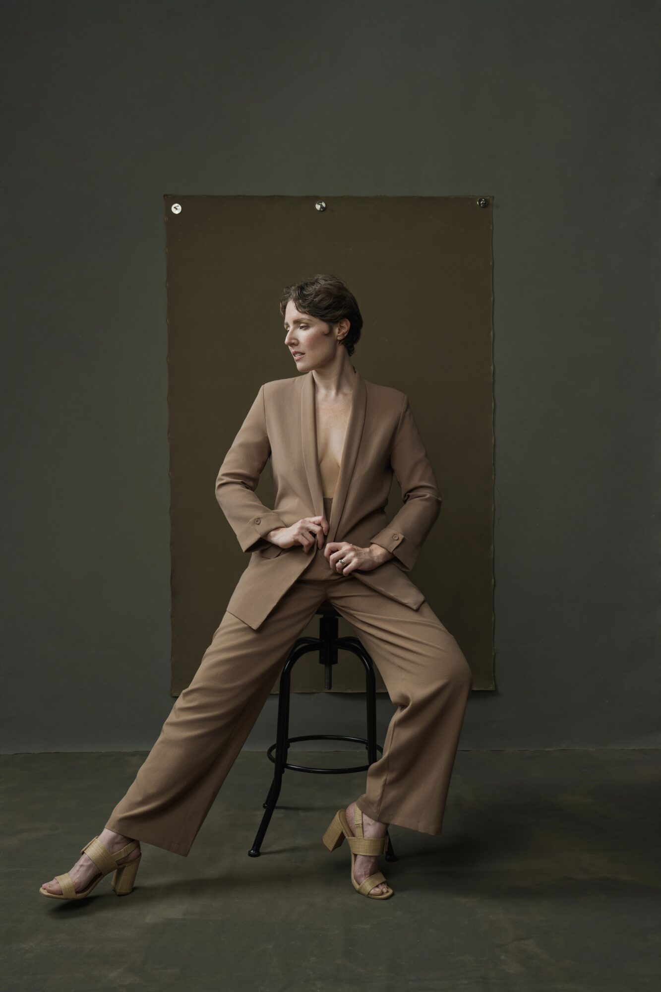 Woman in beige suit sitting on a stool, posing against a dark background.