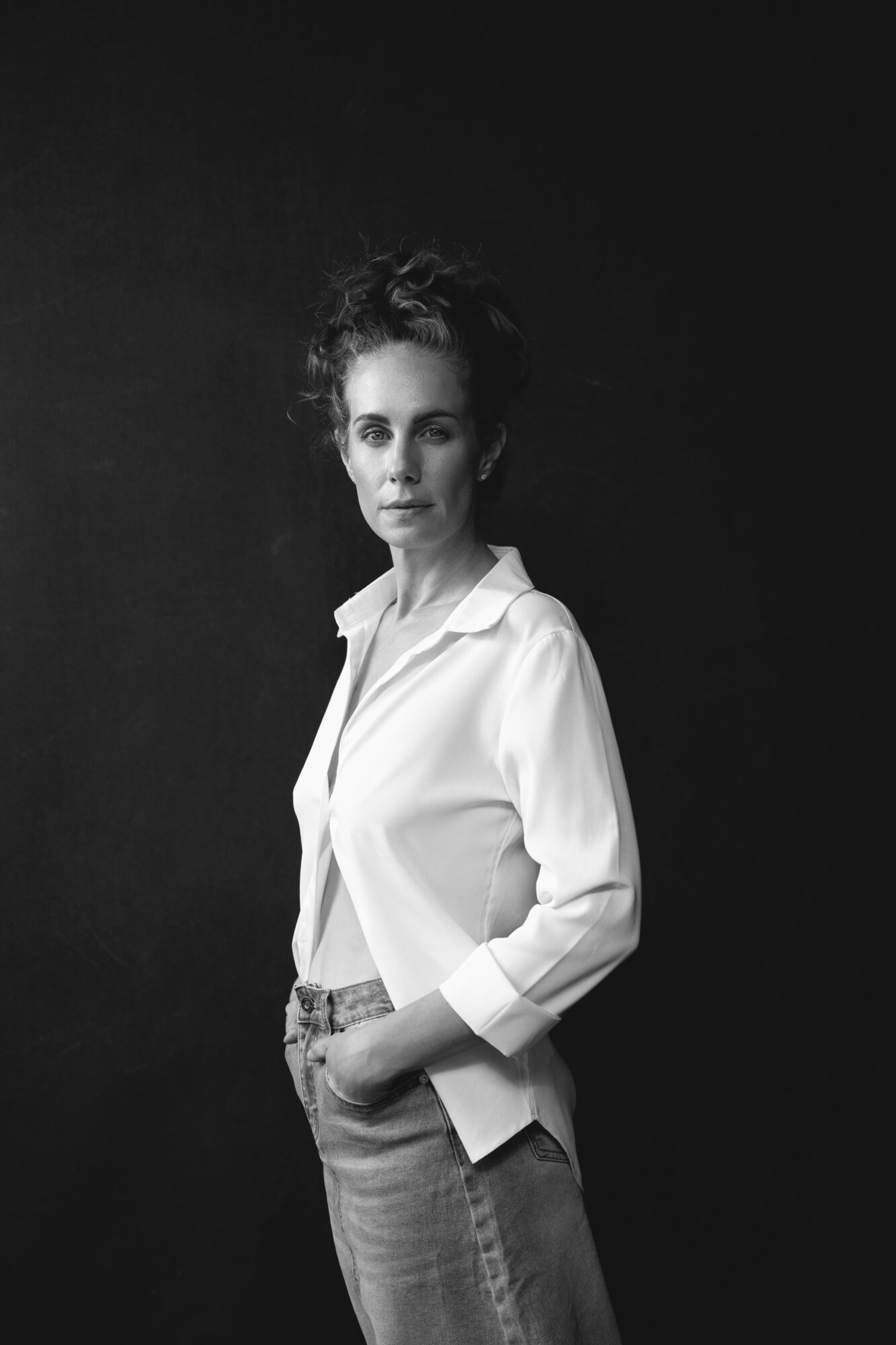 Woman with curly hair in a white shirt and jeans stands against a dark background.