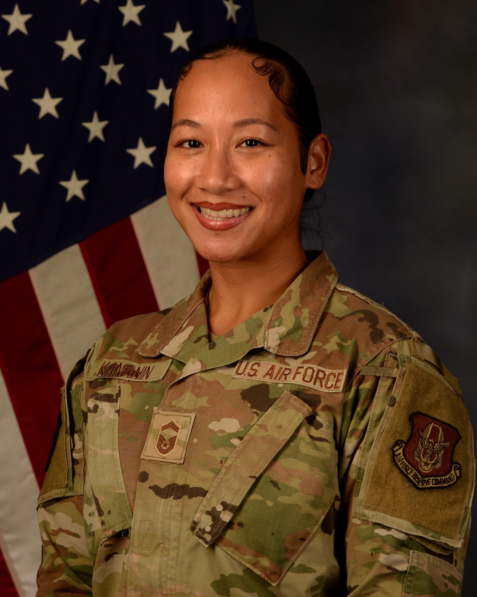 Woman in military uniform smiling, standing in front of an American flag, with dark background.
