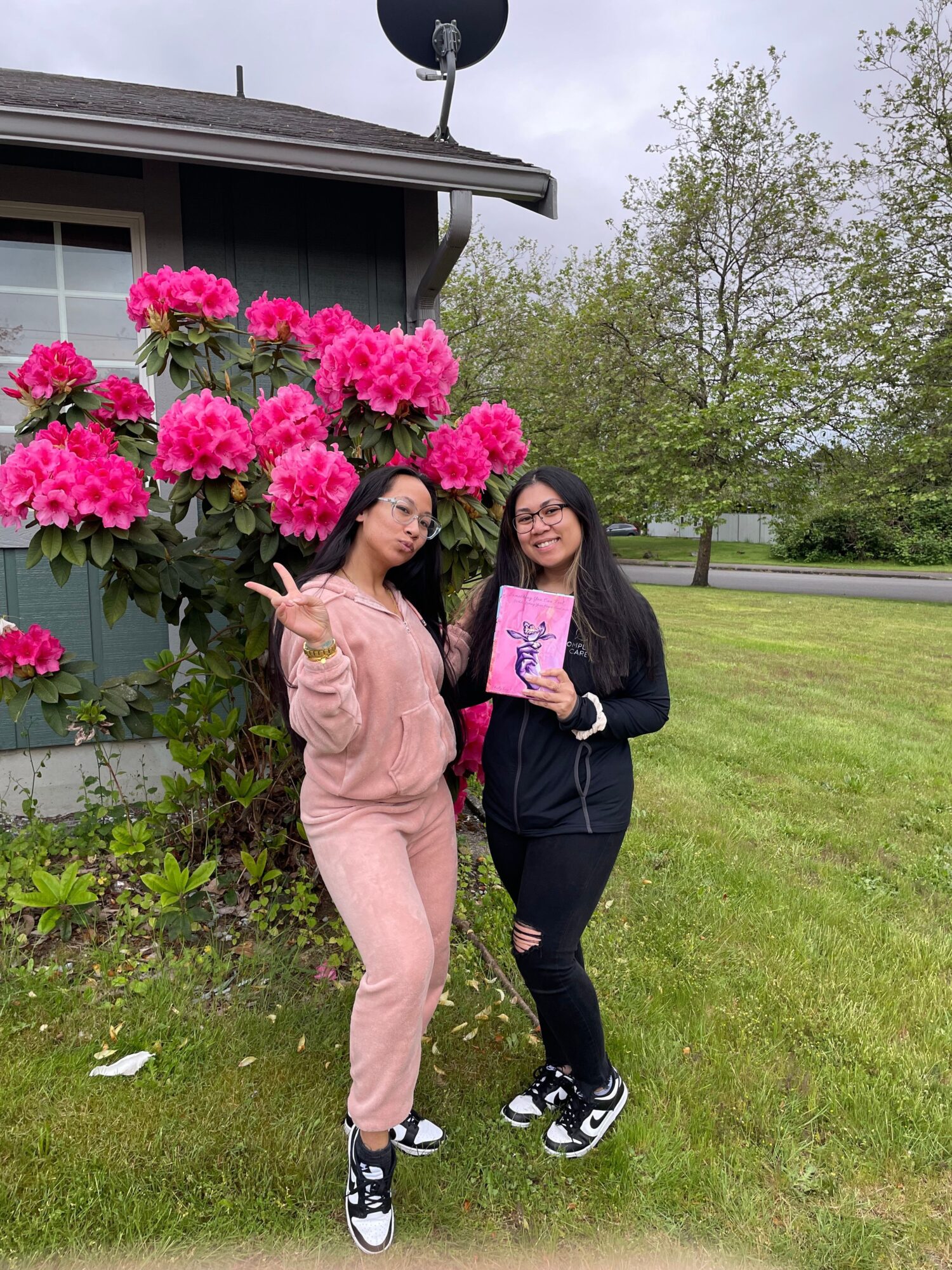 Two women standing outdoors near pink flowers, one holding a pink book, with trees and grass in the background.
