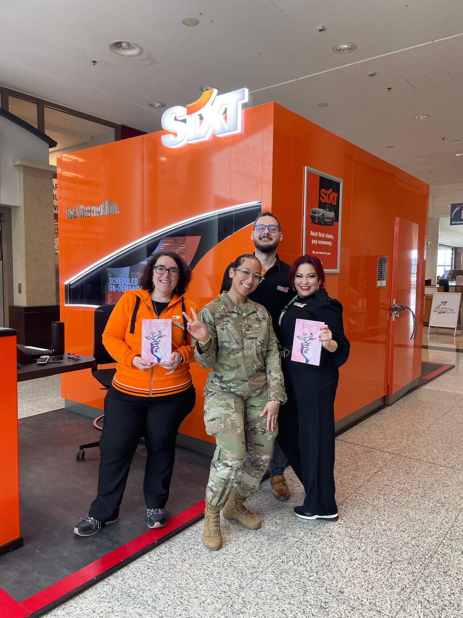 Three people standing in front of an orange wall with a sign, two holding papers, one in military uniform.