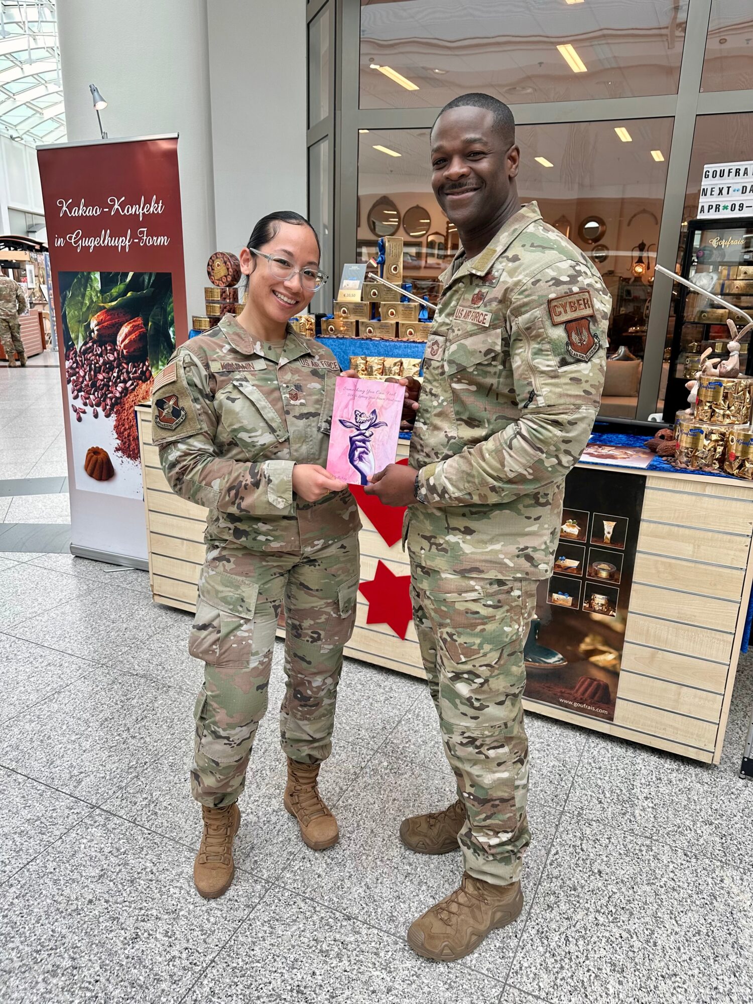Two soldiers in uniform stand inside a shopping mall, holding a pink gift bag together, smiling at the camera.
