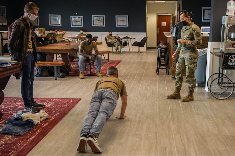 Person doing push-up on floor with military personnel and others in a room, some seated at tables, some standing, with framed pictures on wall.
