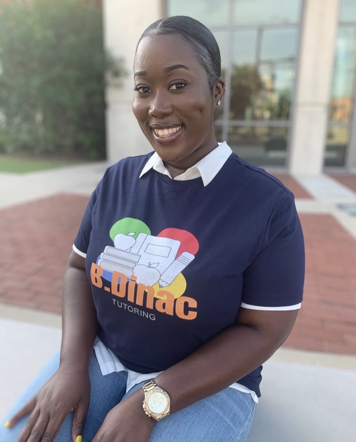 Smiling woman in a navy T-shirt with a logo, standing outdoors with a building in the background.