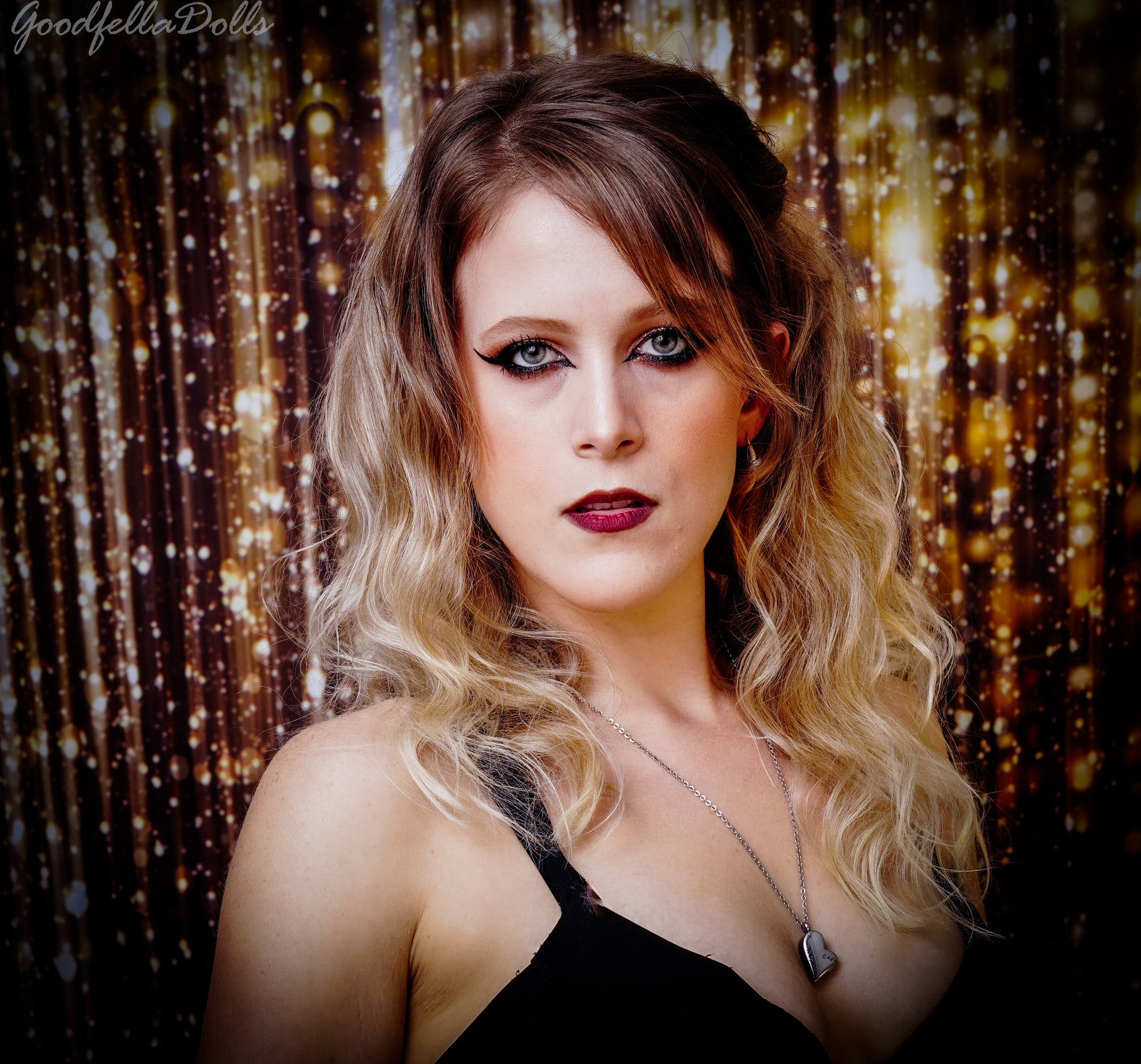 Woman with wavy hair and dark makeup poses in front of a gold, glittery background.