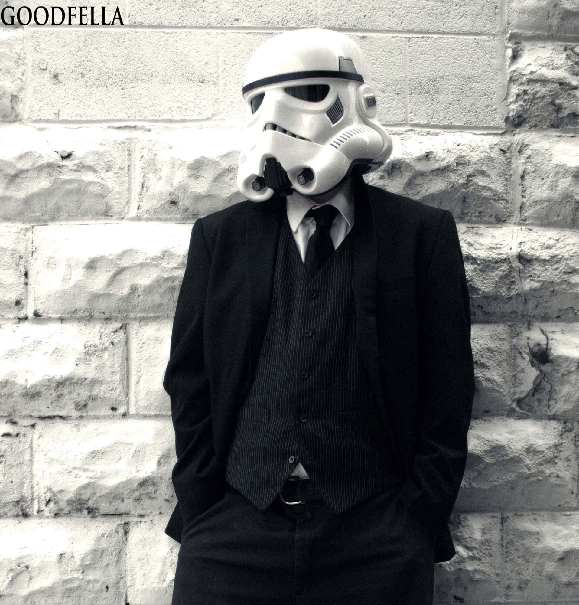 Person in a suit wearing a Stormtrooper helmet, standing against a brick wall, hands in pockets.