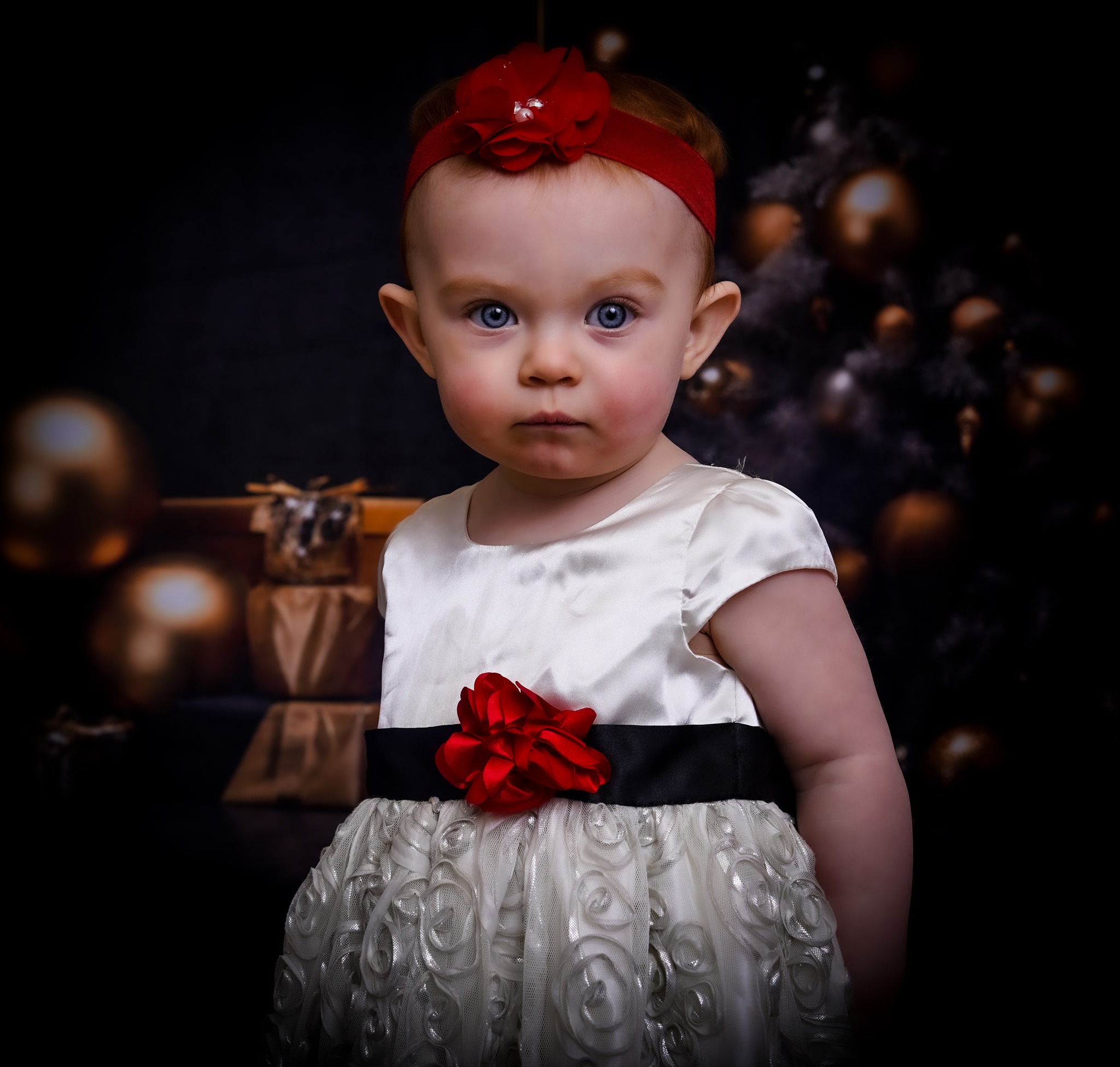 Young girl with a red headband and dress, standing in front of a dark background with Christmas decorations.