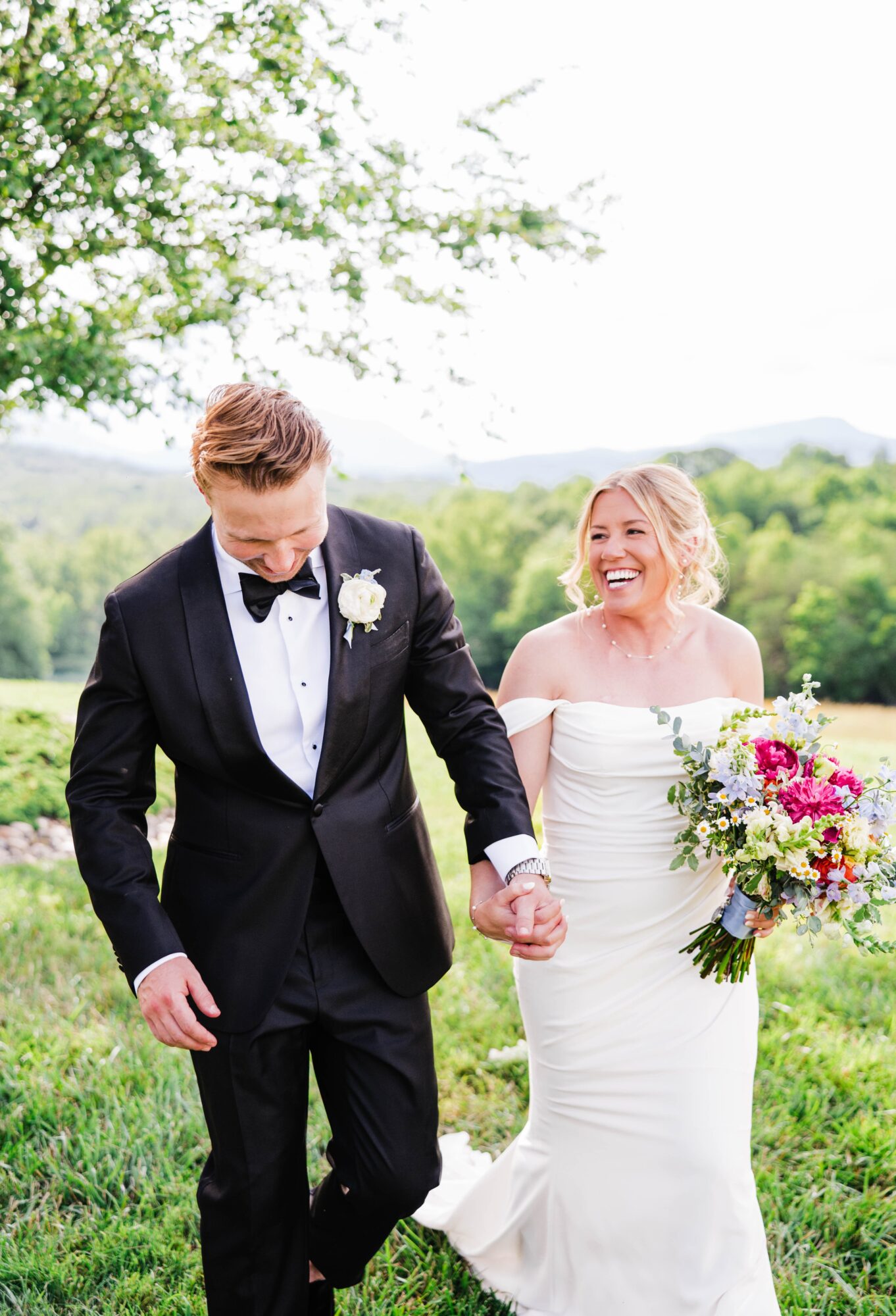 A bride and groom holding hands outdoors, smiling, with trees and a landscape in the background.
