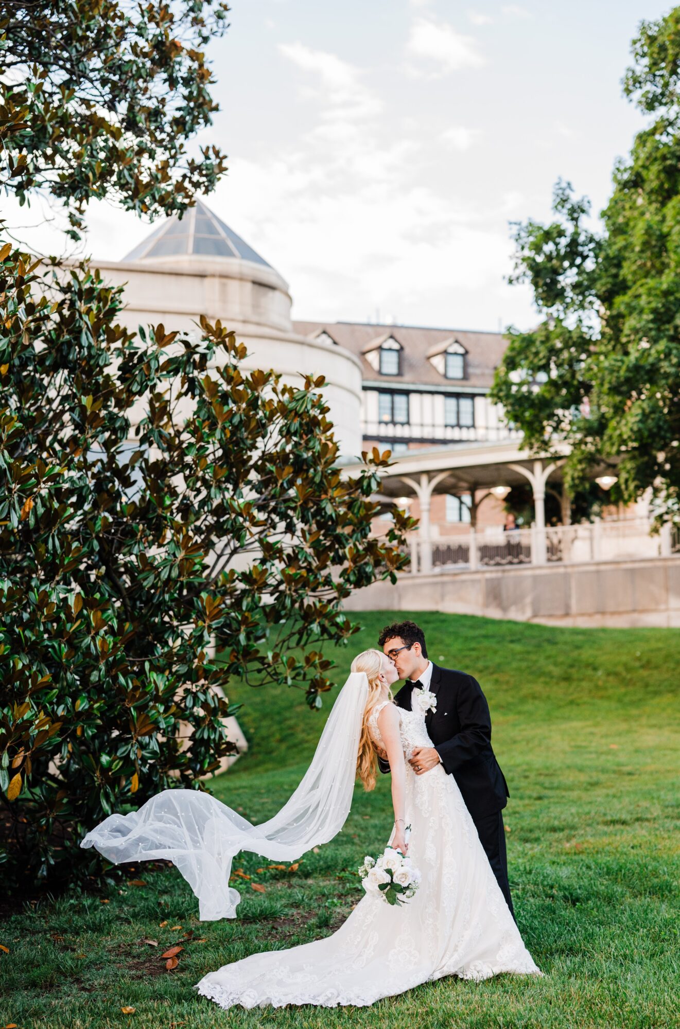 Bride and groom kissing outdoors on grass with a large building and trees in background.