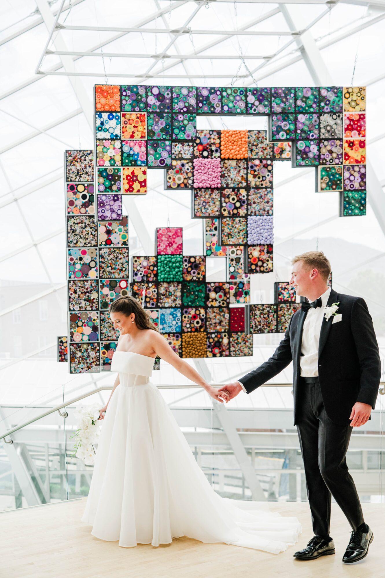 Bride and groom holding hands in front of a colorful quilt display in a modern glass building.