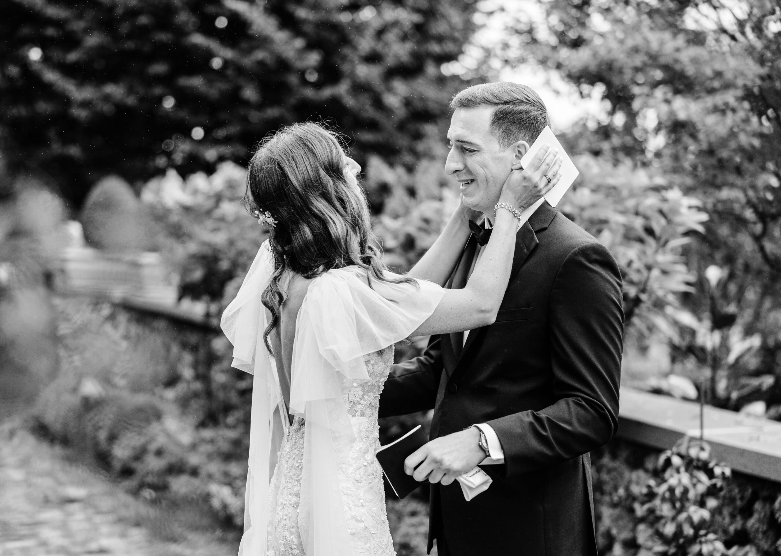 A woman adjusts a man's tie outdoors, both smiling, with trees and a fence in the background.