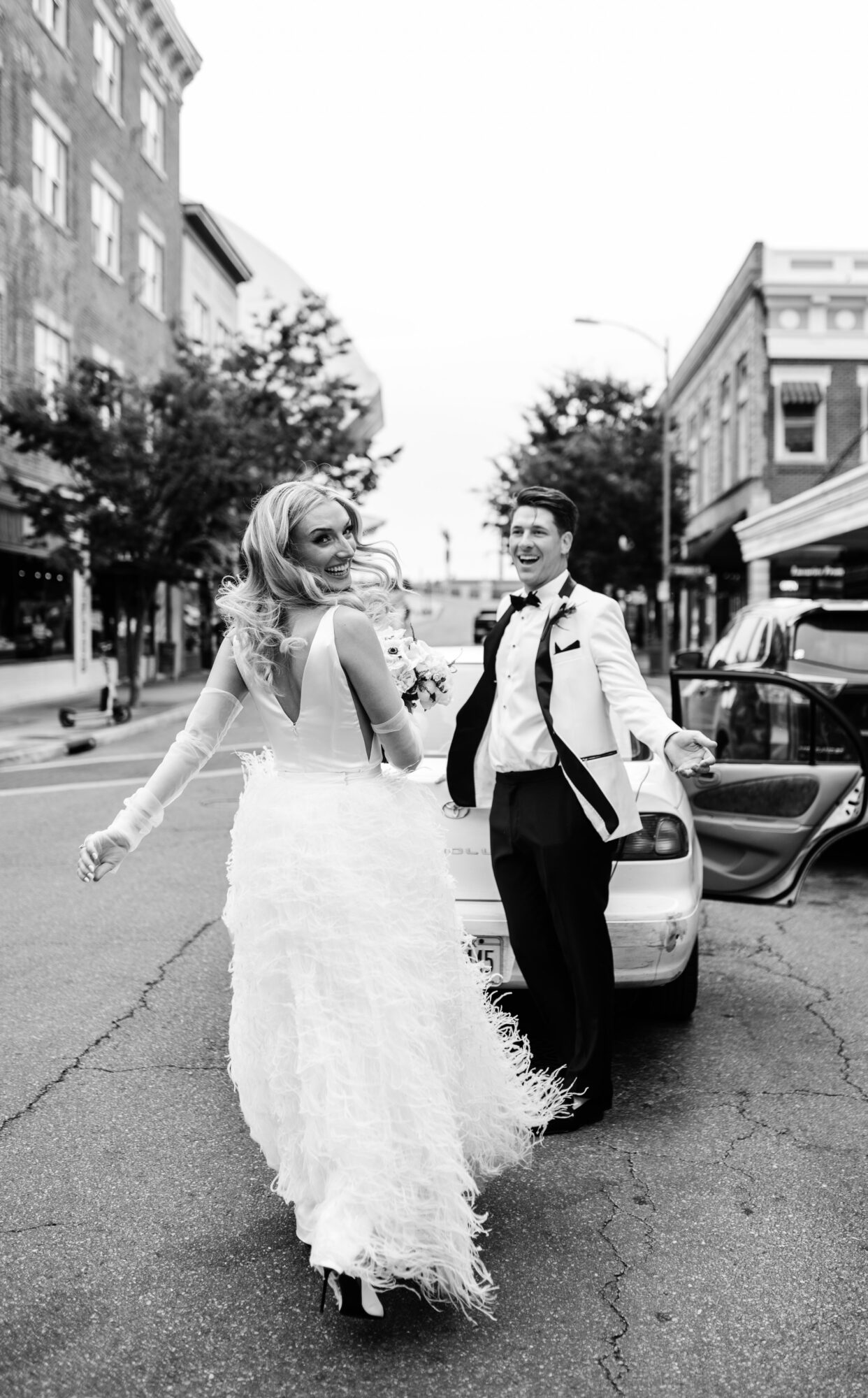 A woman in a wedding dress and a man in a suit stand on a city street, smiling and holding hands.
