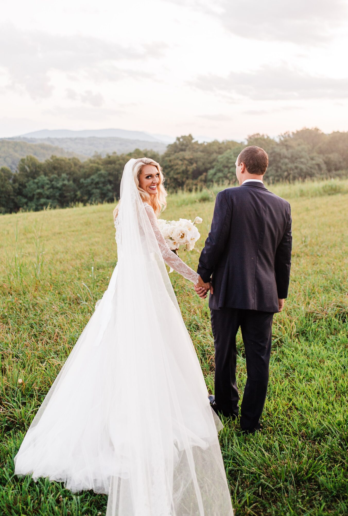 Bride and groom holding hands in a grassy field, bride in a white wedding dress, groom in a dark suit, landscape background.