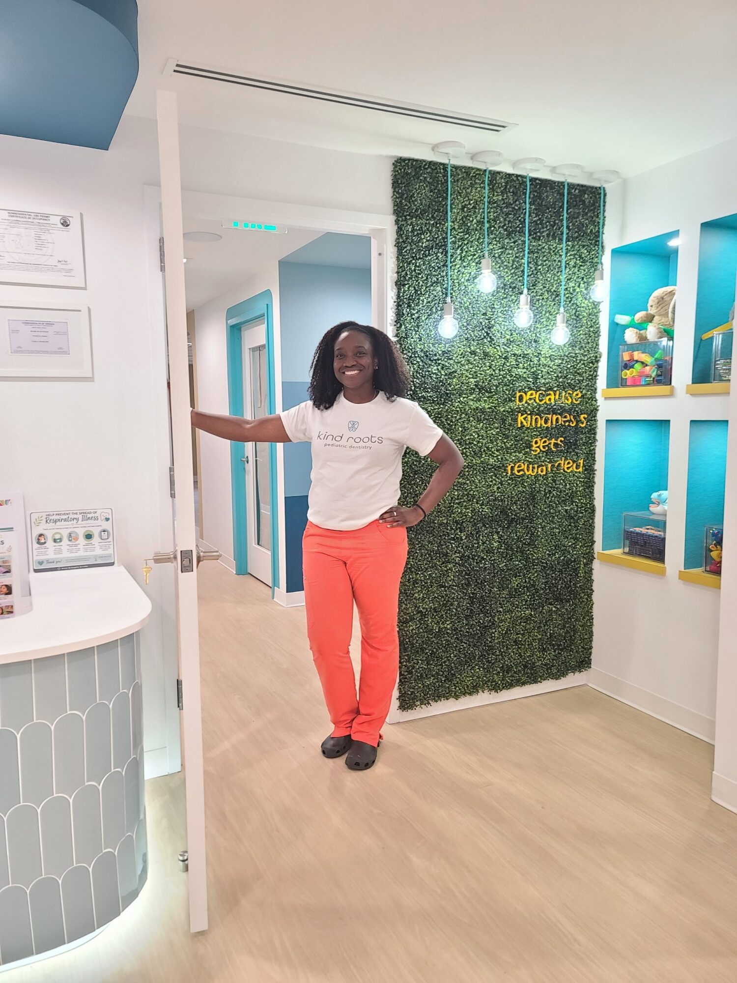 Woman standing in a brightly lit room with a green wall, shelves, and hanging lights, smiling with one hand on her hip.