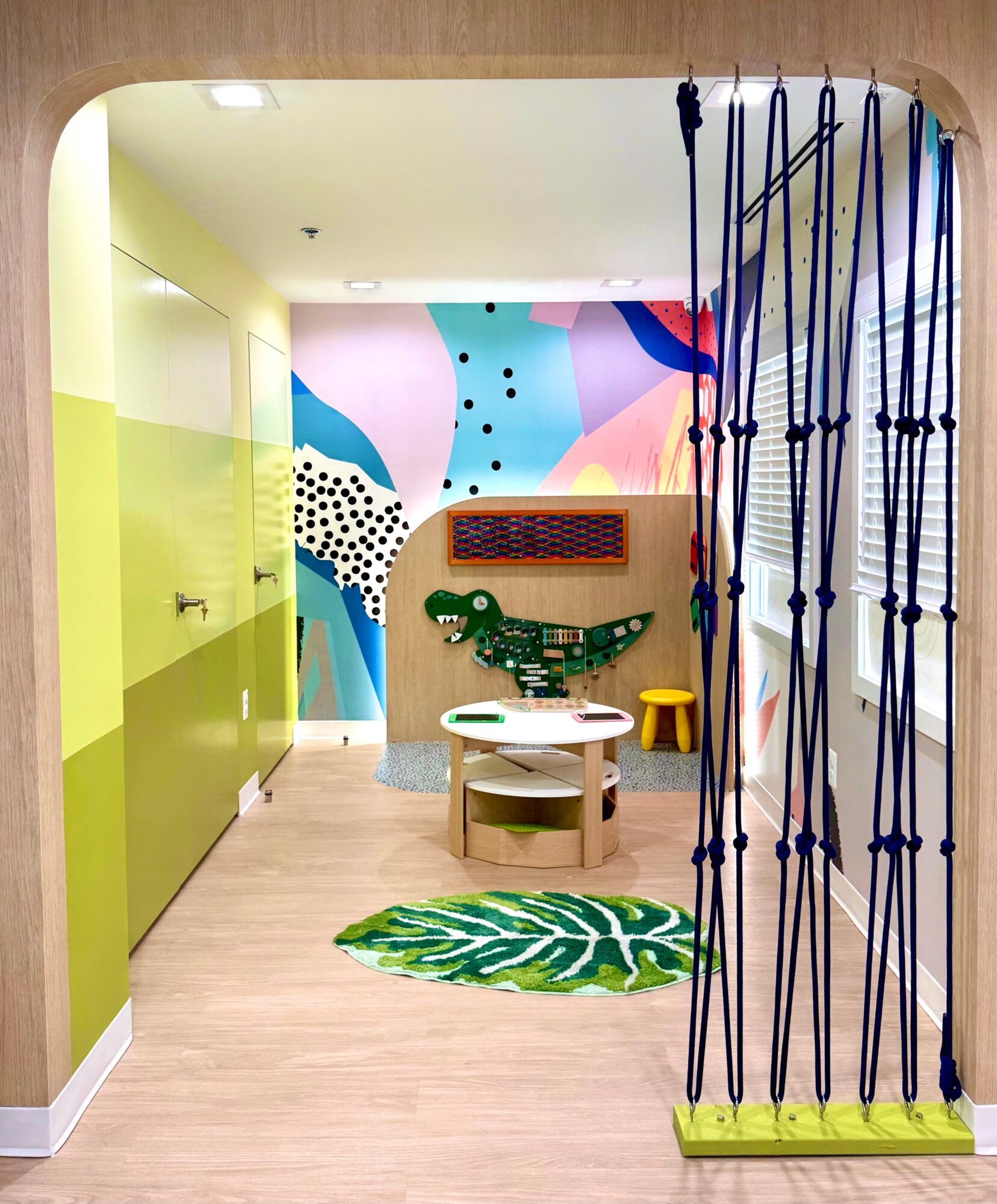 Children's playroom with colorful wall mural, small table and chairs, green leaf rug, and a yellow stool, viewed from doorway.